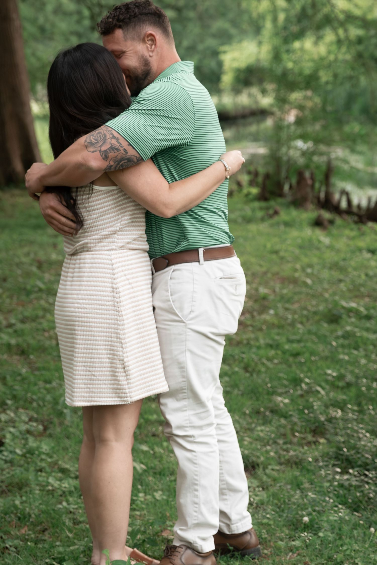 Couple embracing outdoors in a lush green park, man in green polo and white pants hugging woman in striped dress. Cedartown, GA