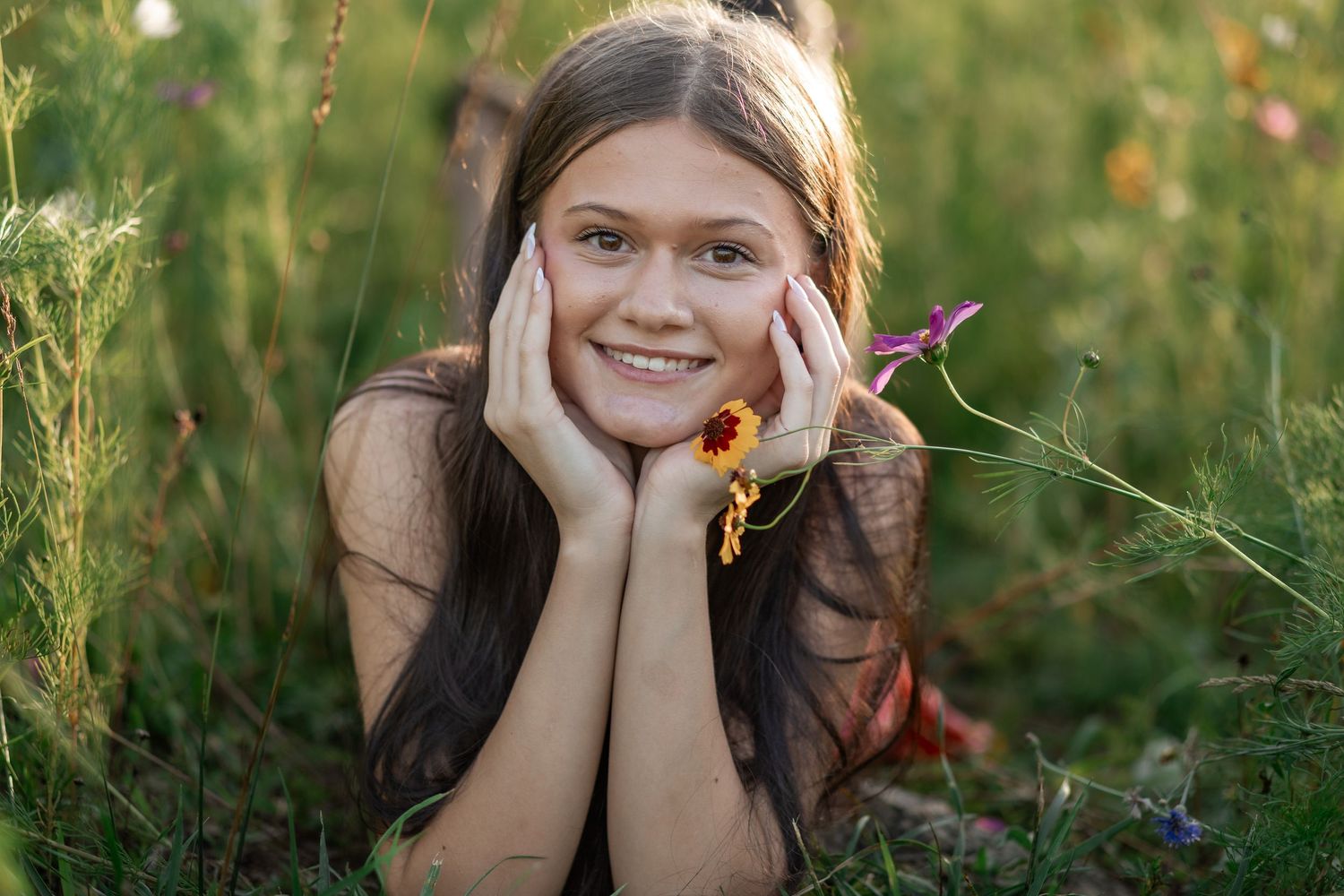 Smiling young girl lying in a wildflower meadow, resting her chin on her hands, holding a colorful flower.