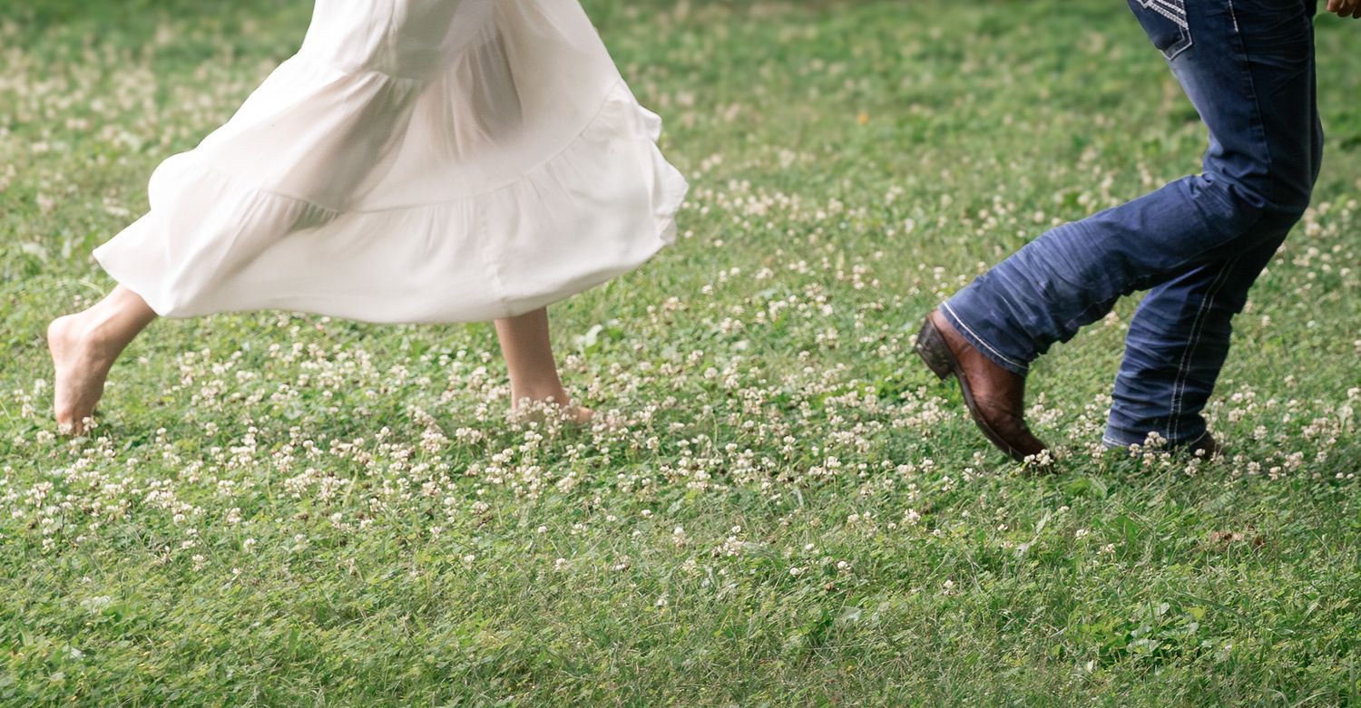 Two people walking barefoot and in boots on a green grass field with white clover flowers. Cedartown, GA