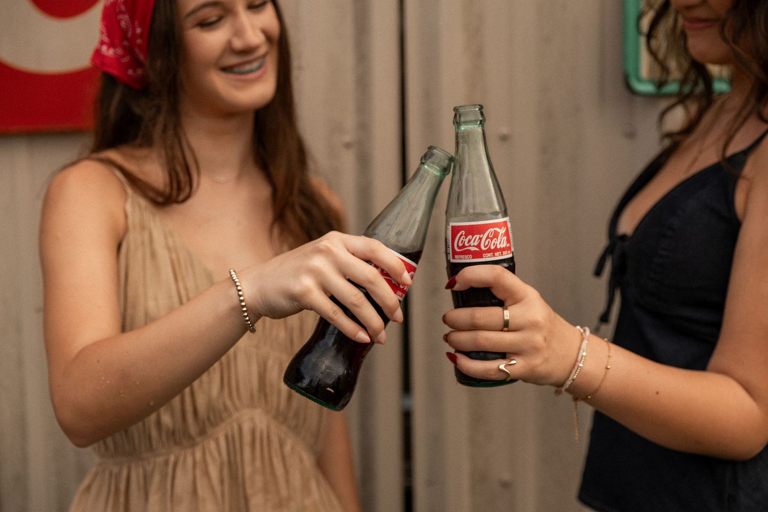 Two young women clinking glass Coca-Cola bottles together in a celebratory toast at a casual gathering.