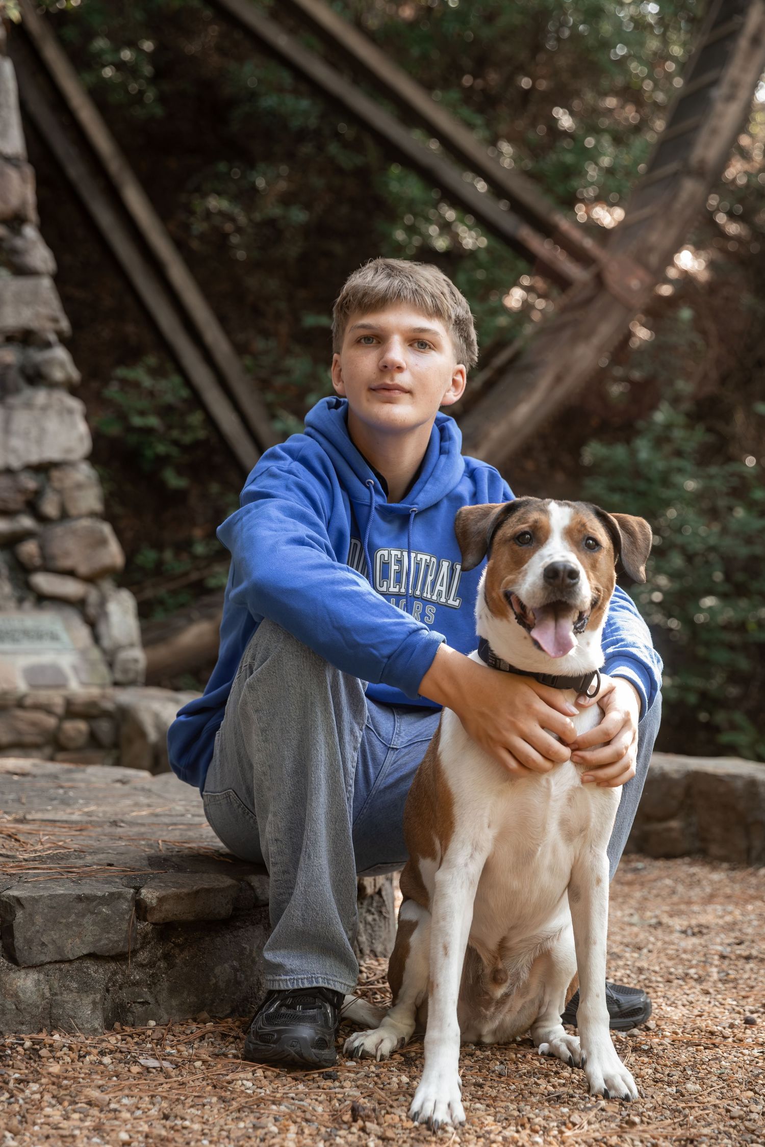 Teen boy in blue Central hoodie sits with brown and white dog outdoors near stone wall and trees.
