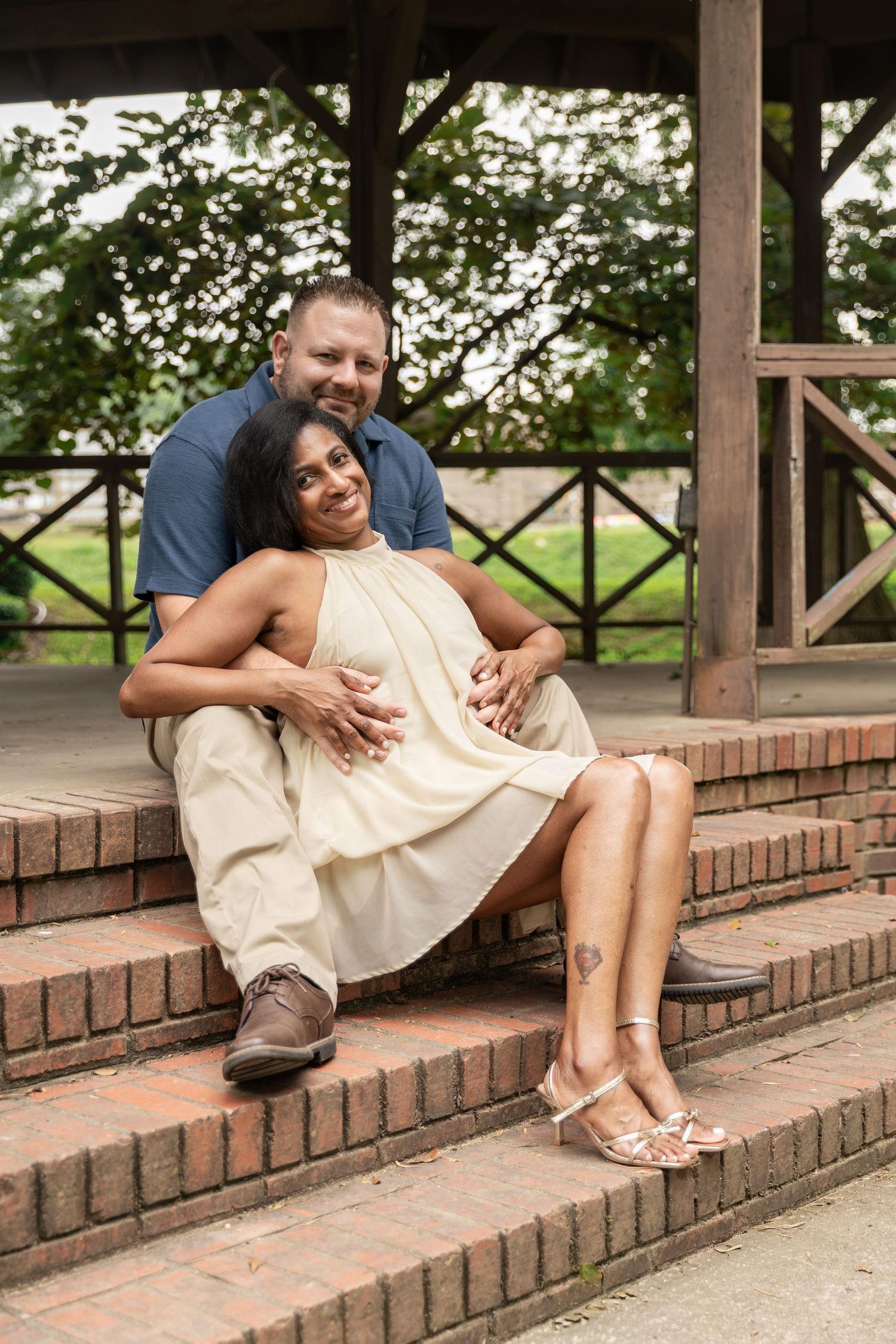 Couple sitting on brick steps in a gazebo, man embracing woman in cream dress, smiling outdoors. Rockmart, GA