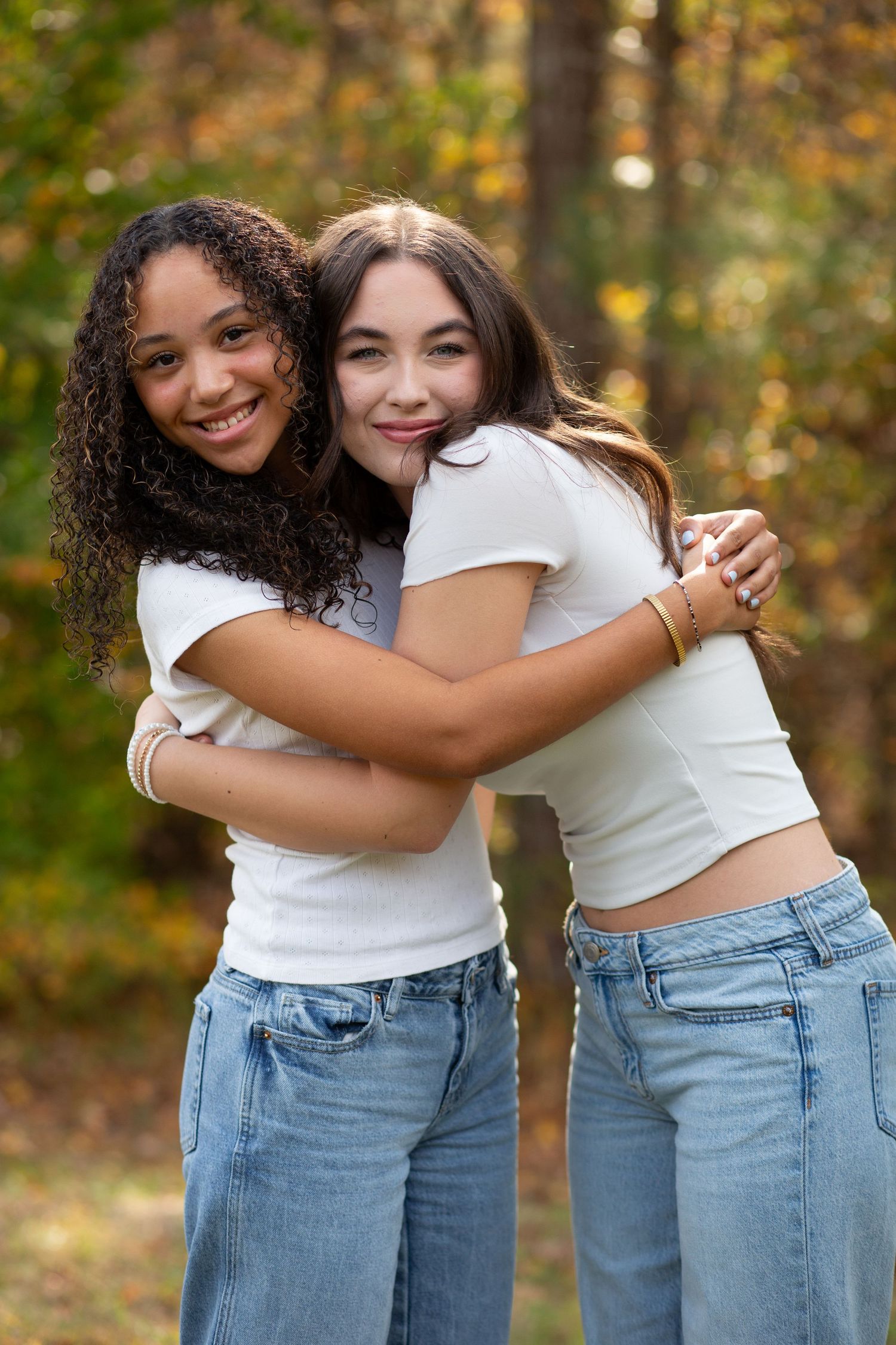 Two teenage girls hugging and smiling outdoors in white t-shirts and jeans with autumn foliage in the background.