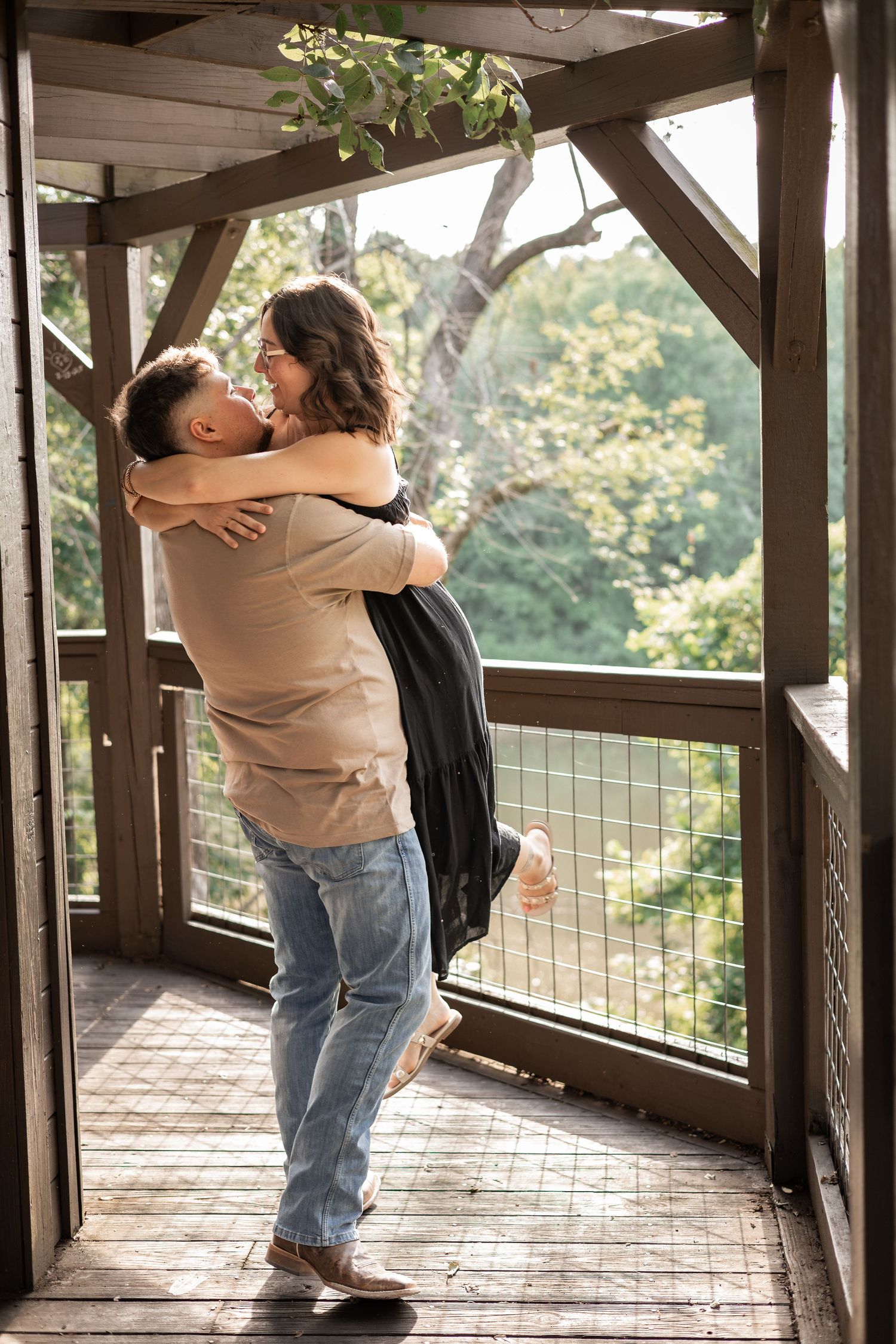 A couple embraces joyfully on a wooden deck, man lifting woman, surrounded by lush green trees in the background.