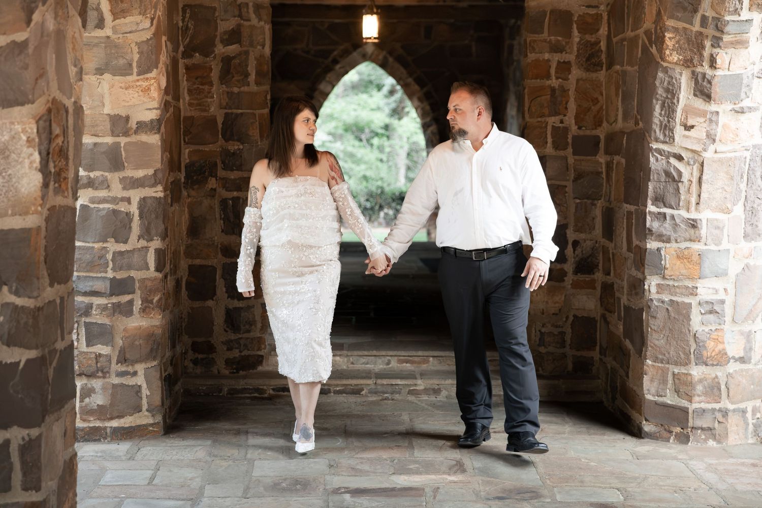 Couple holding hands and smiling at each other under a stone archway, woman in white lace dress, man in white shirt. Berry College, Rome, GA