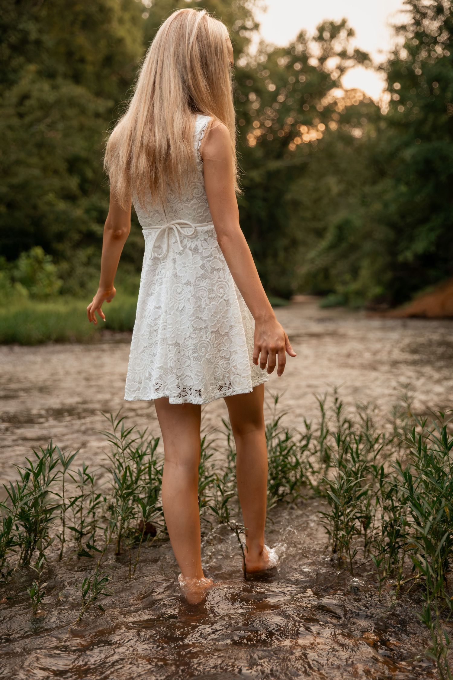 Blonde woman in white lace dress walking barefoot through a sandy creek bed surrounded by green plants.