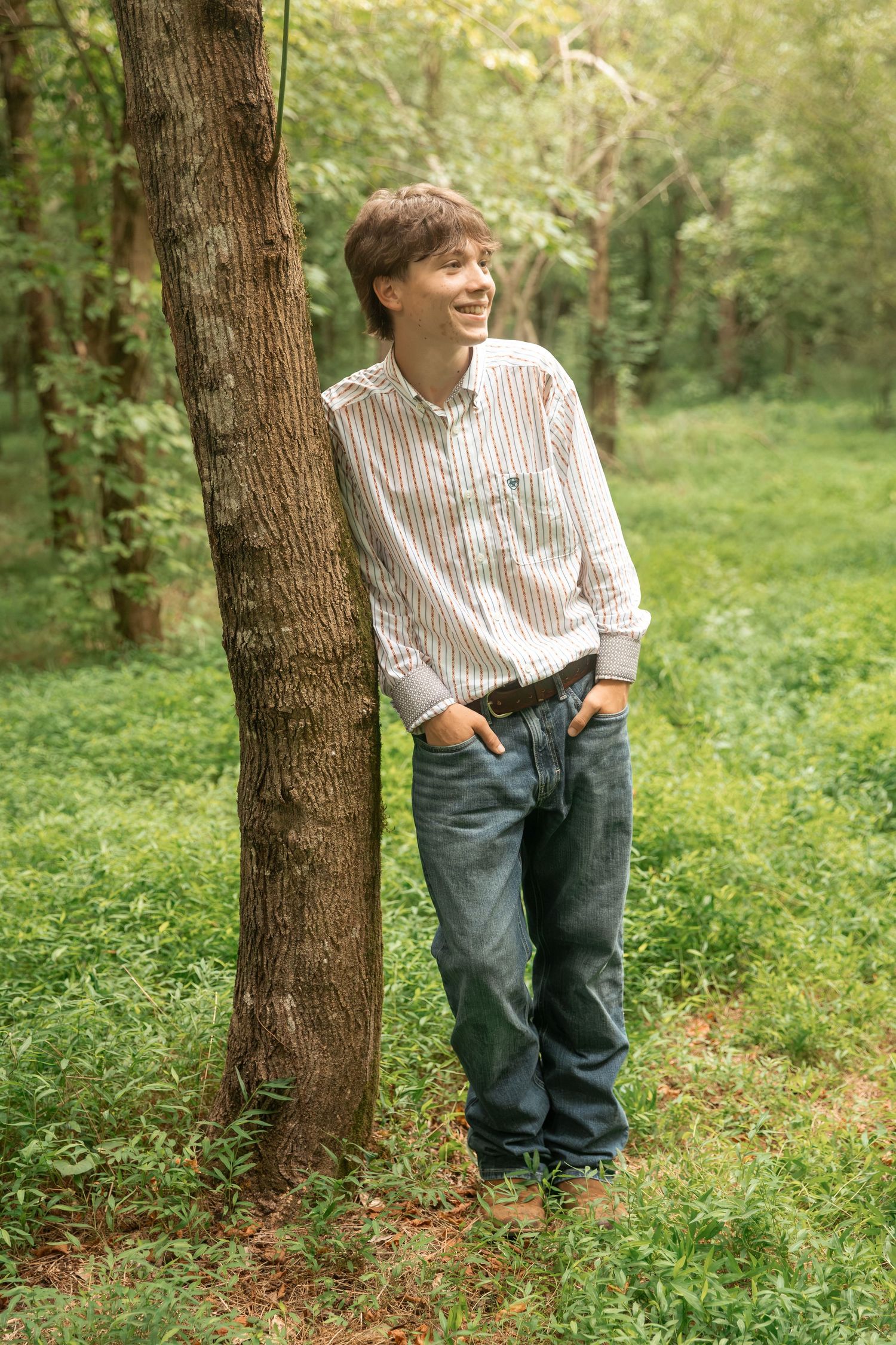 Happy teenage boy laughing while leaning on a tree in a green woodland setting, wearing jeans and a striped shirt.