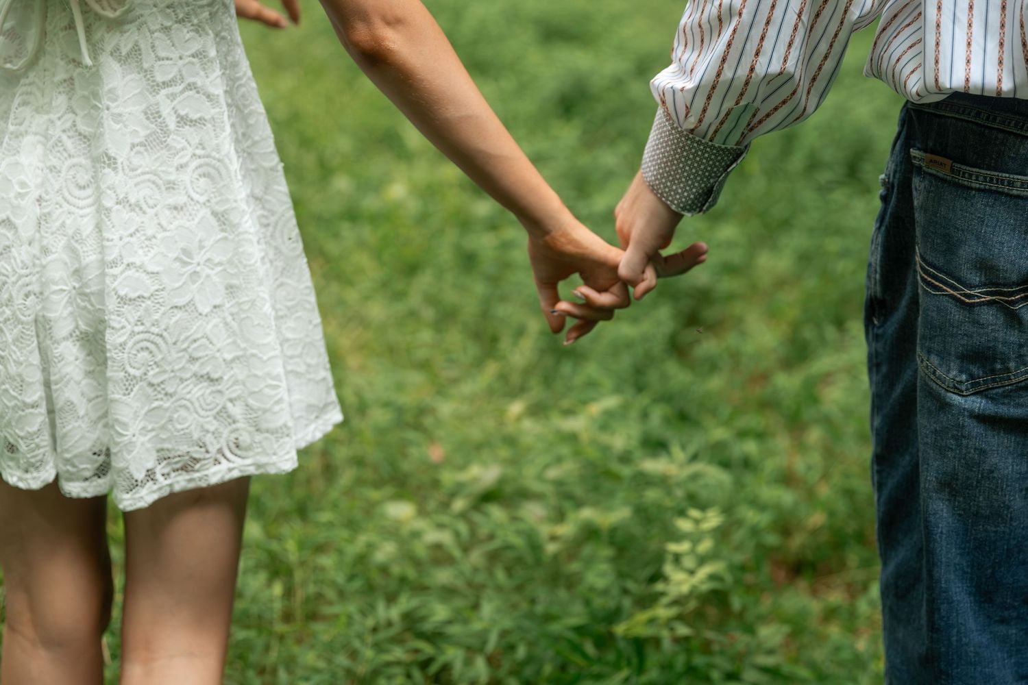Couple holding hands while walking through a lush green field, woman in white lace dress and man in plaid shirt. Rome, GA