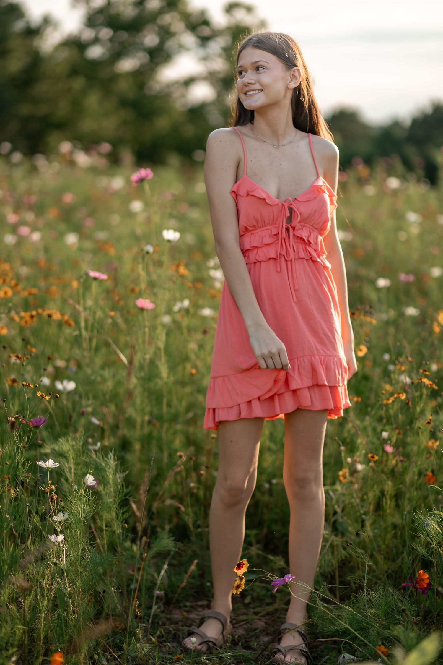 Young woman in coral ruffle dress smiling in a wildflower meadow at golden hour.