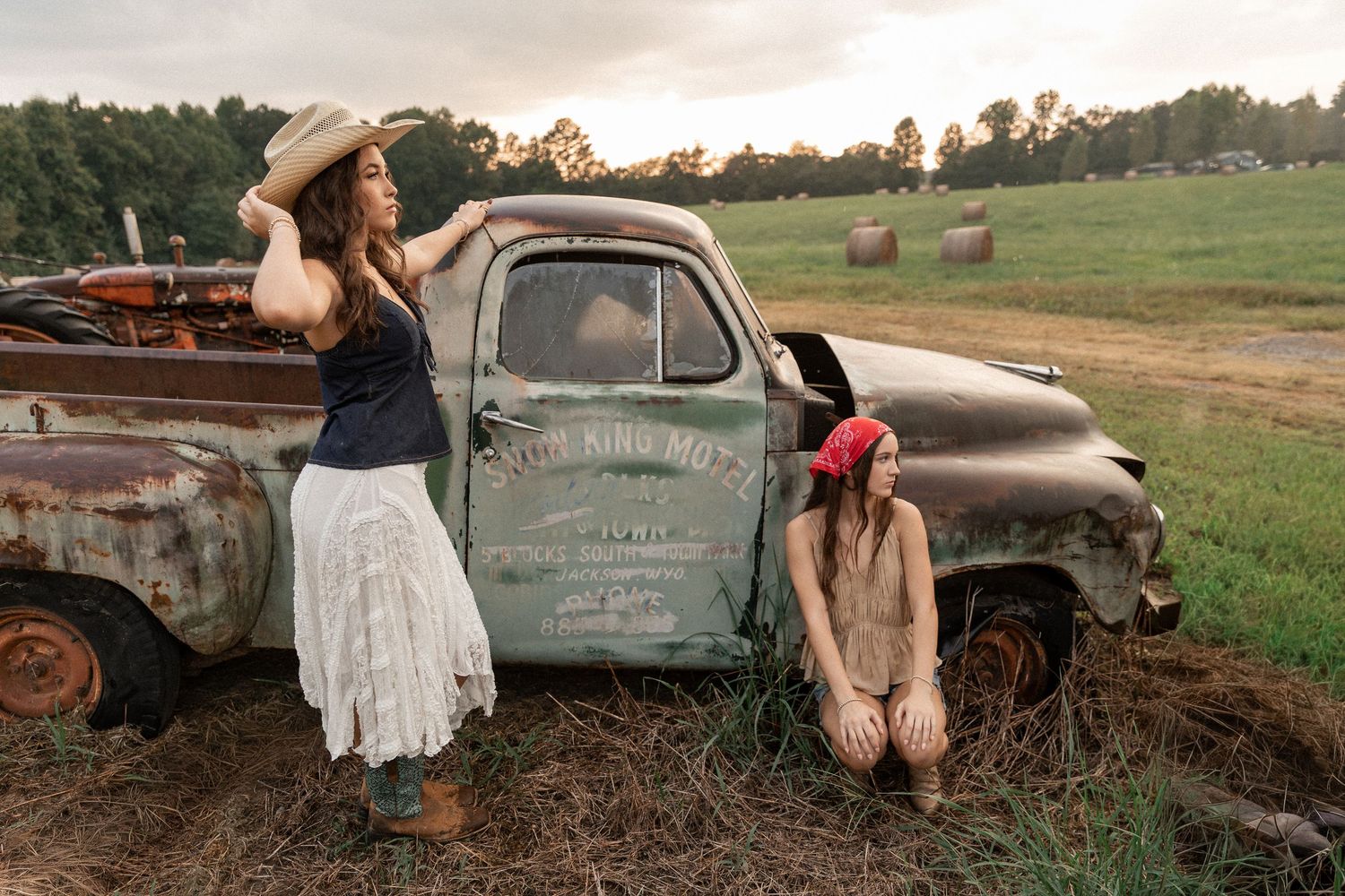 Two women pose by a rustic vintage truck in a green field with hay bales, wearing country-style outfits.