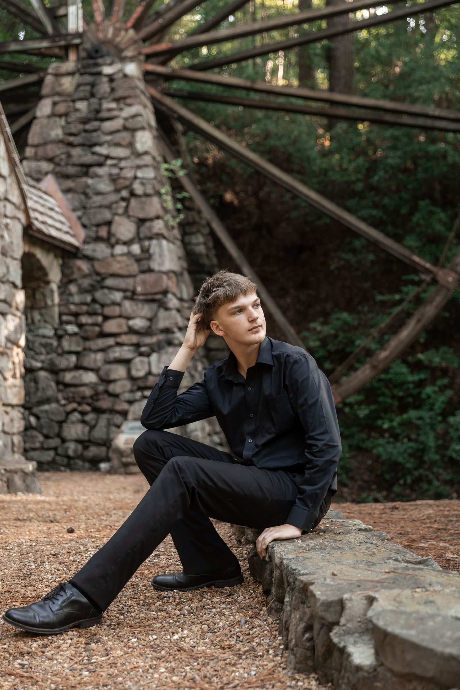 Young man in all-black outfit sitting on stone wall near rustic windmill and forest backdrop.