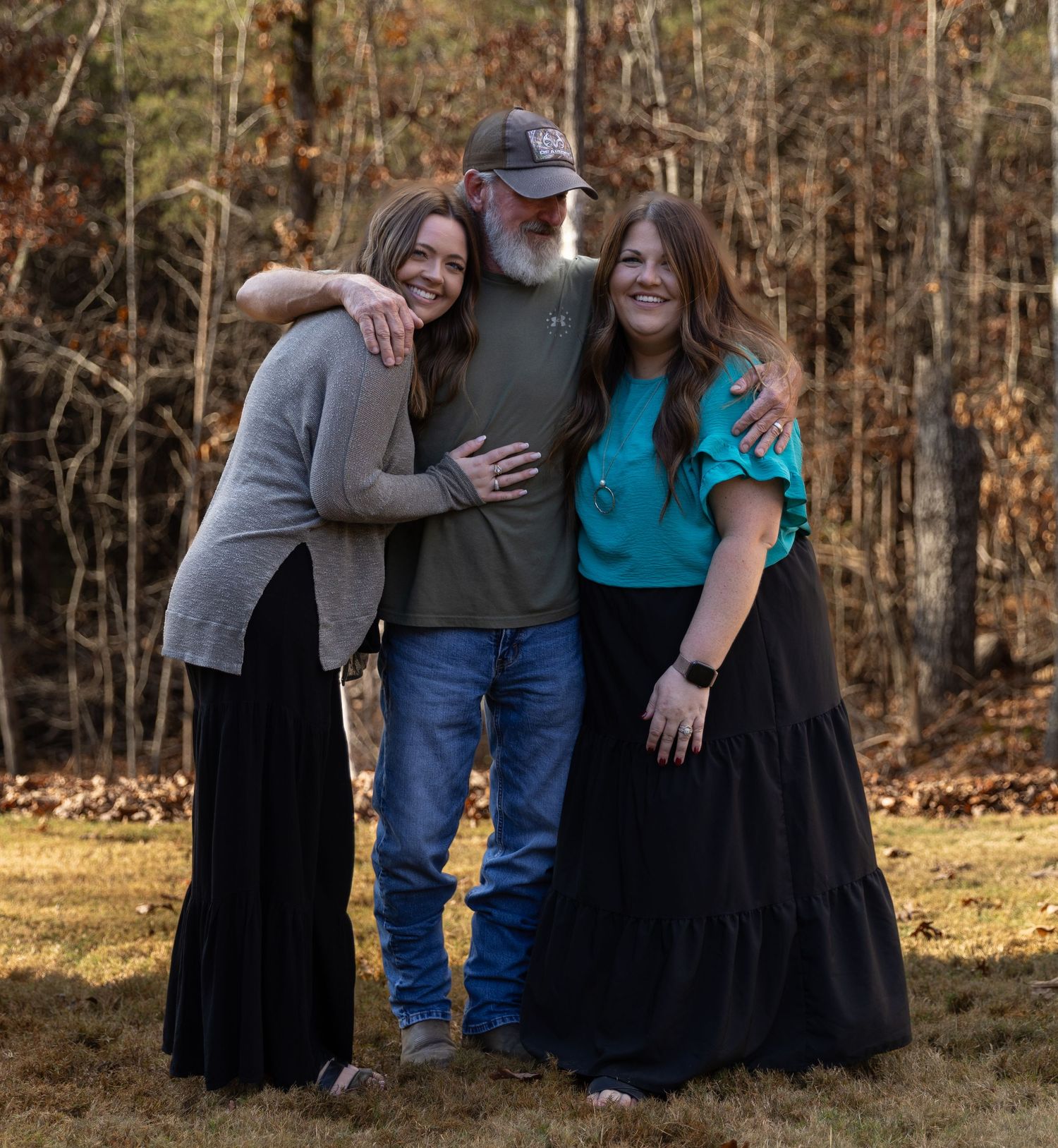 Three smiling people posing together outdoors in a wooded autumn setting, wearing casual clothing.