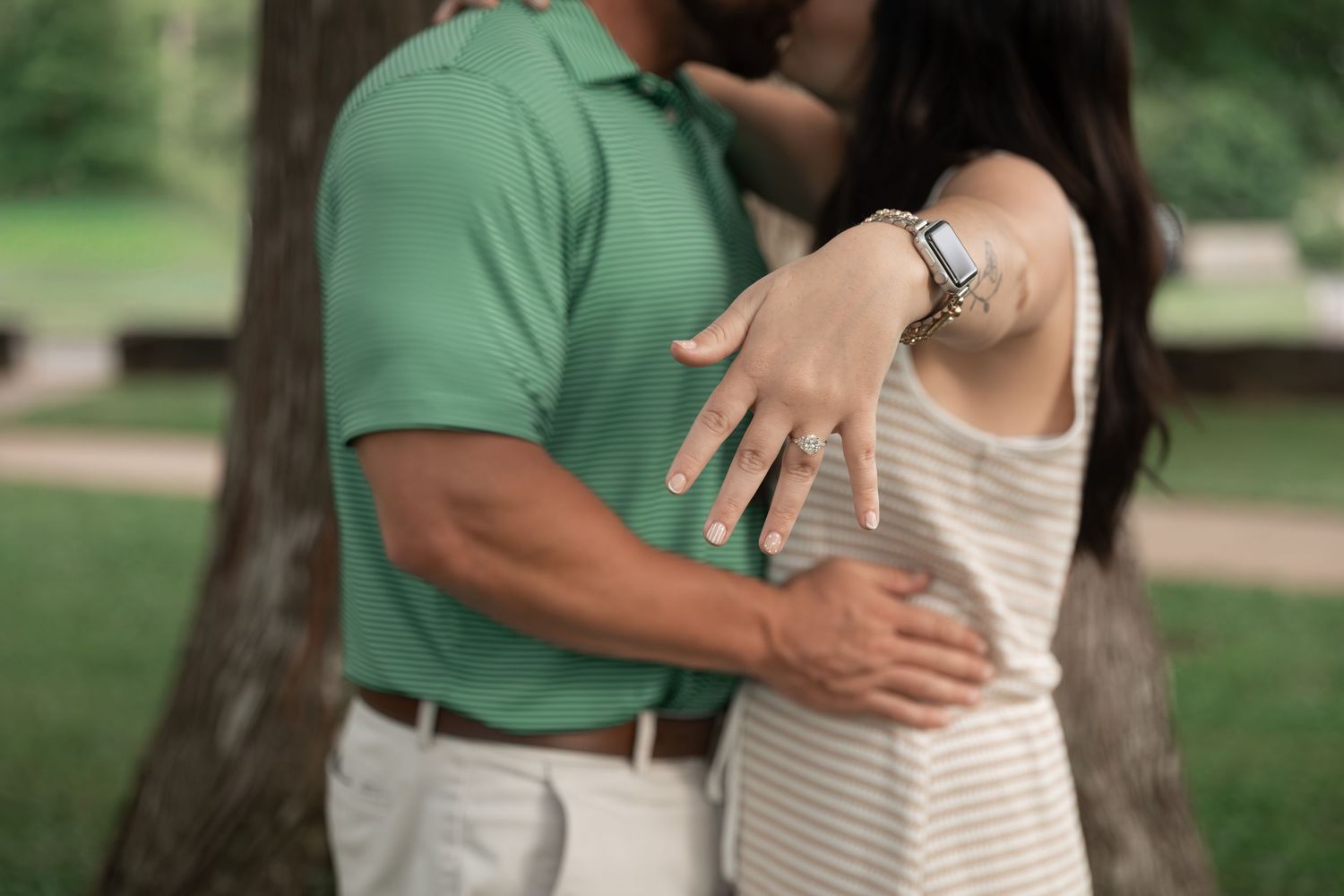 Couple embracing and kissing outdoors, woman showing off engagement ring on her hand, wearing a striped dress and watch.