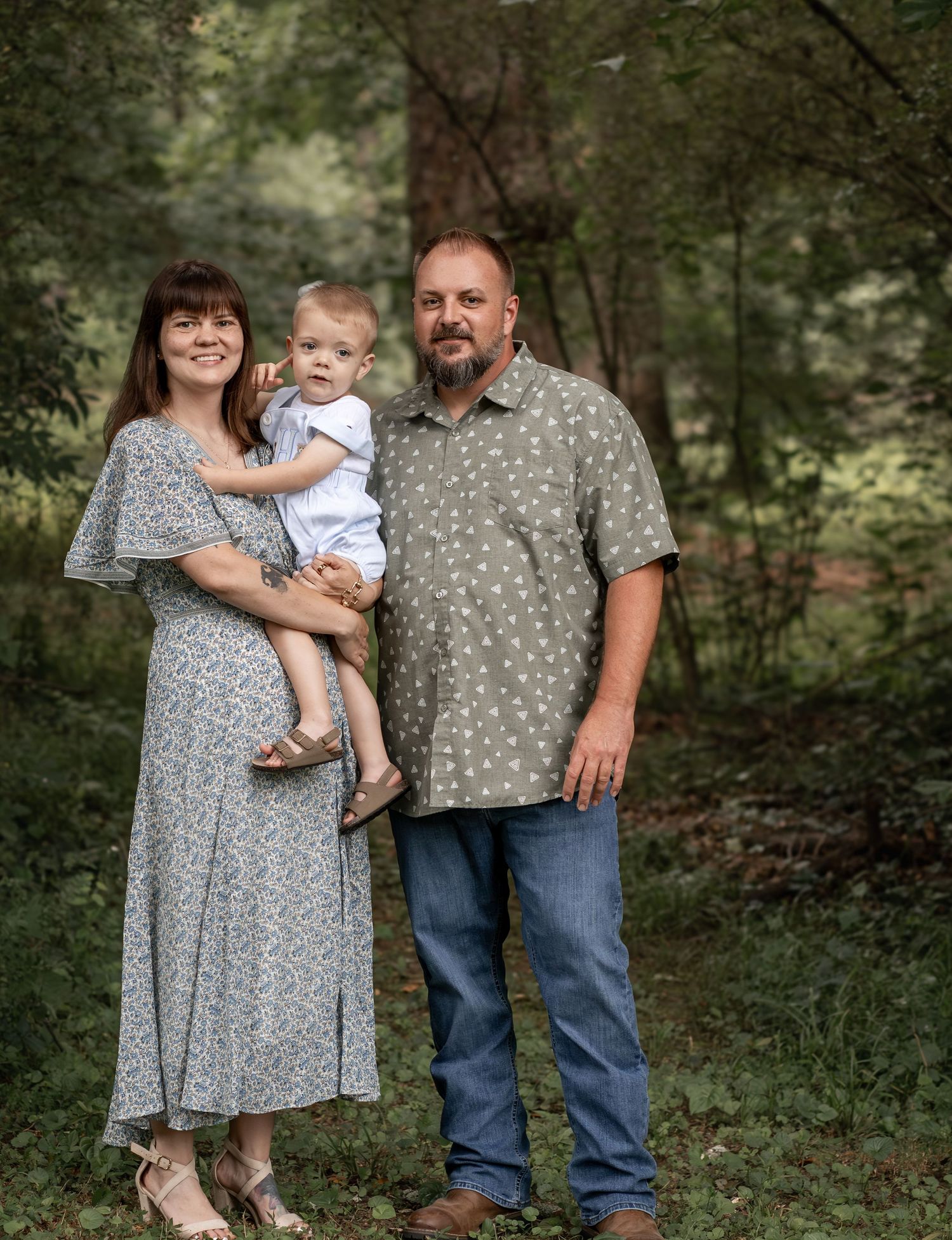 A smiling family of three poses outdoors in a lush green forest setting, with parents holding their young toddler.