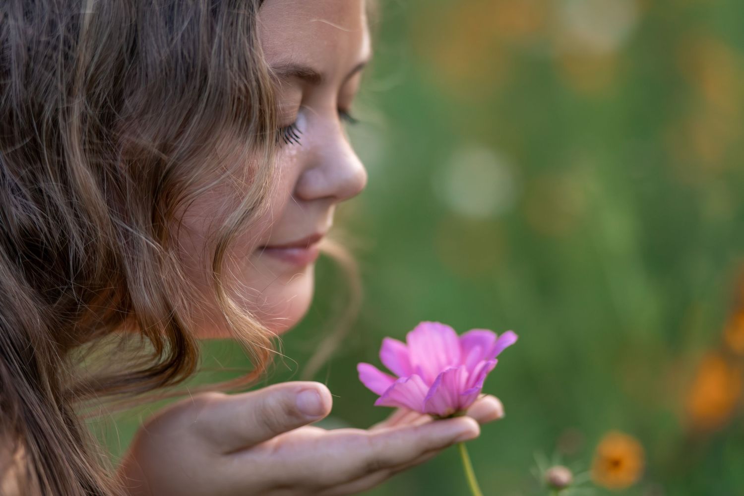 A young girl with closed eyes smells a pink flower held in her hands in a green field.