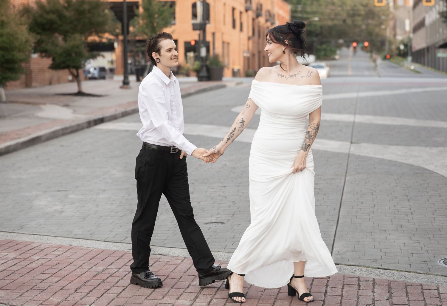 Bride and groom hold hands walking on brick street, she wears white off-shoulder gown with tattoos, he wears white shirt.