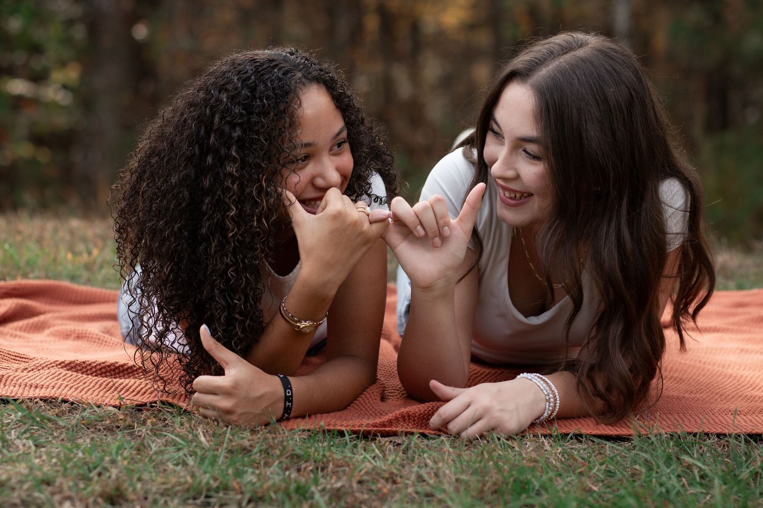 Two young women laughing and bonding while lying on an orange blanket outdoors in a wooded setting.