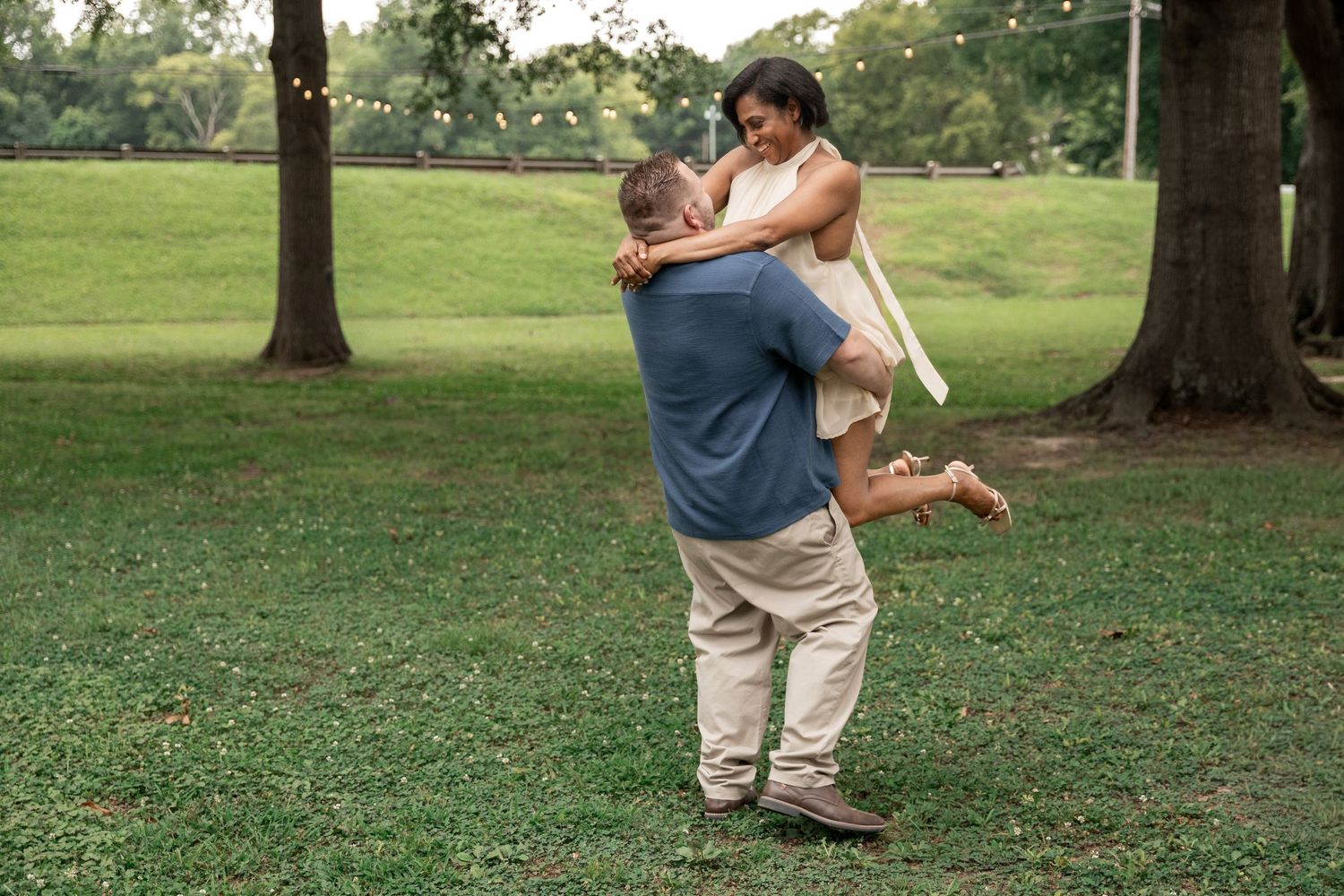 A man joyfully lifts a woman in a sunny green park, sharing a romantic moment surrounded by tall trees. Rockmart, GA
