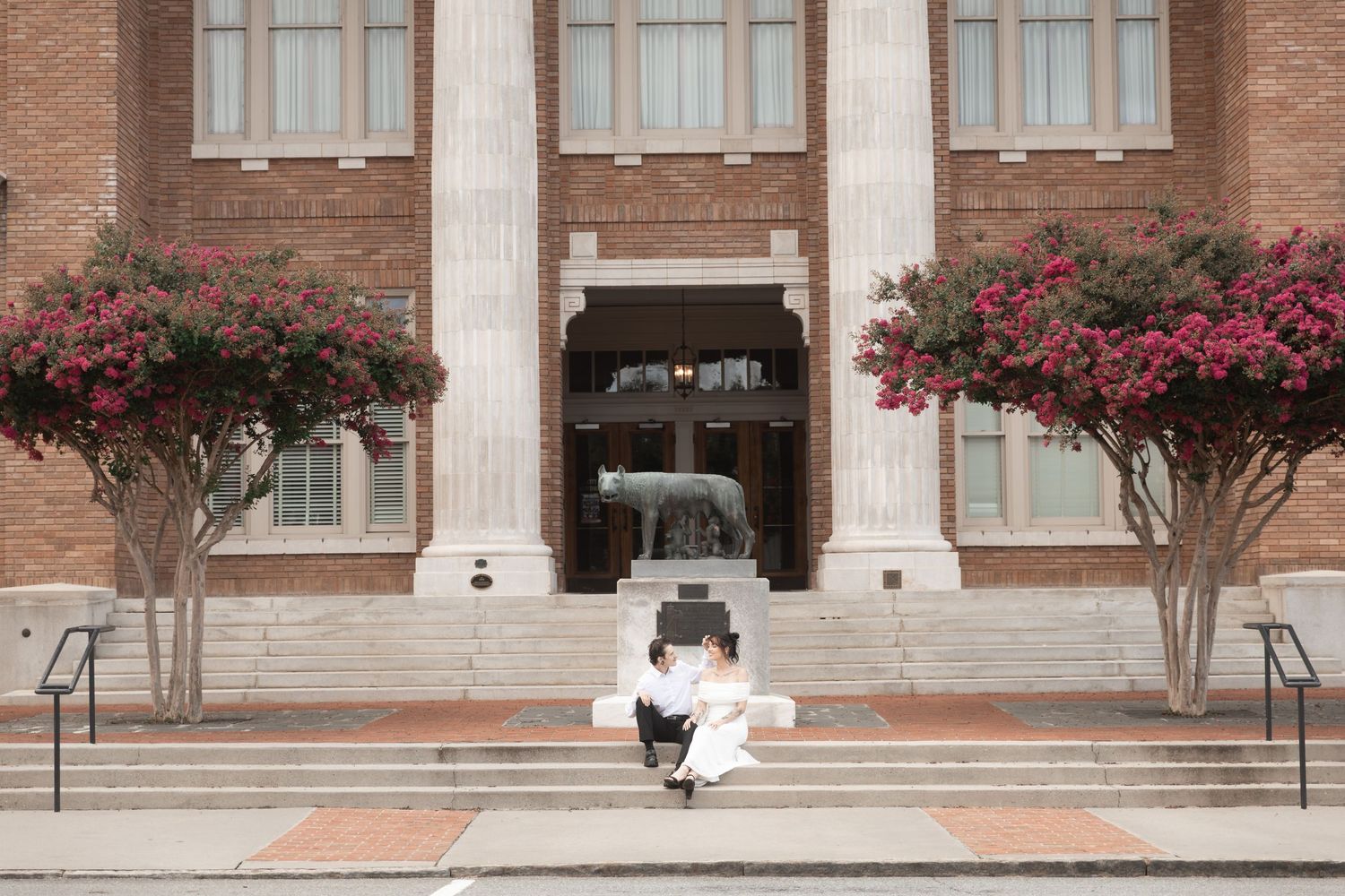 Couple embracing on steps of brick building with white columns and blooming red crepe myrtle trees.  Downtown Rome, GA