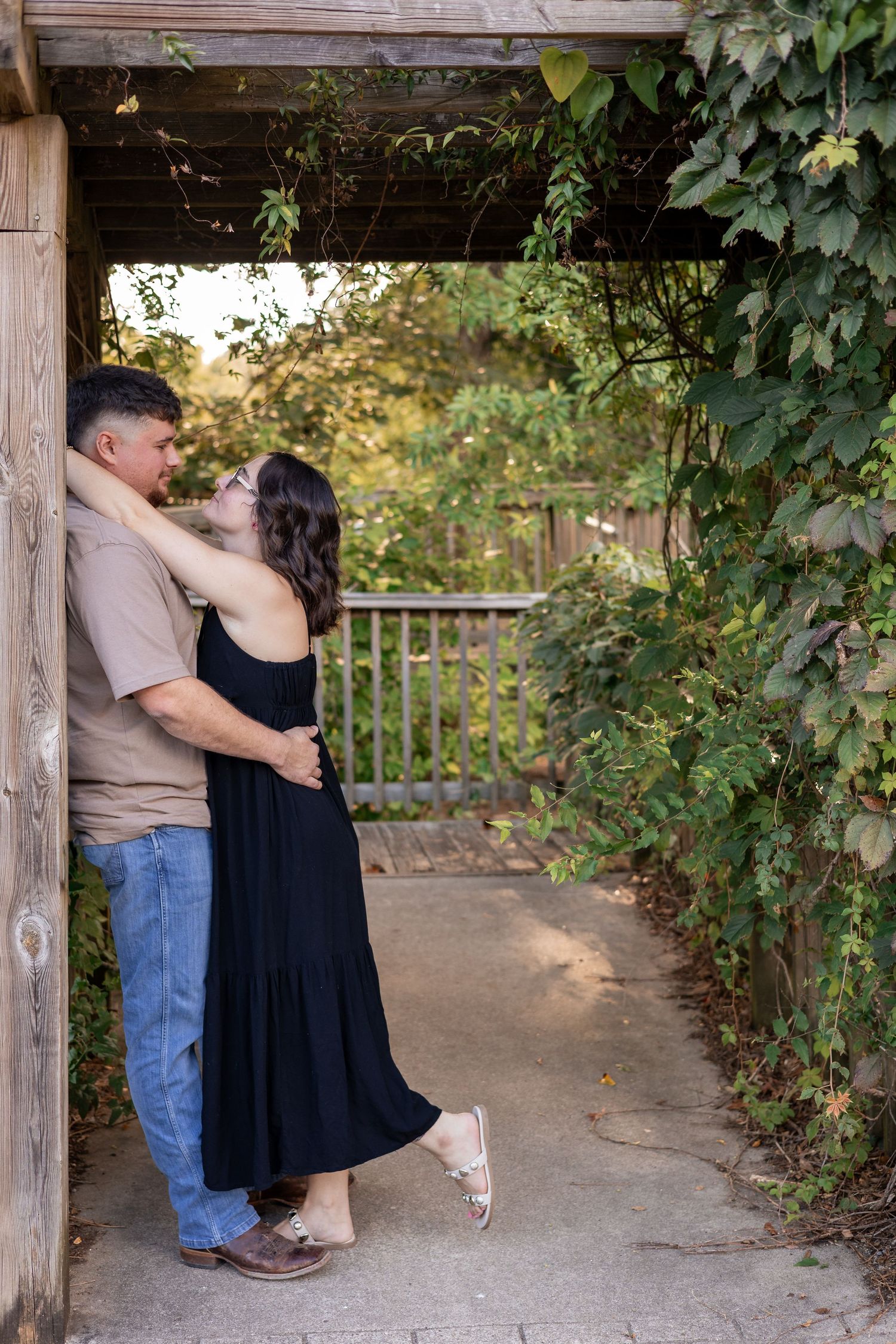 Couple embracing romantically under a wooden arbor covered in lush green vines in an outdoor garden setting.