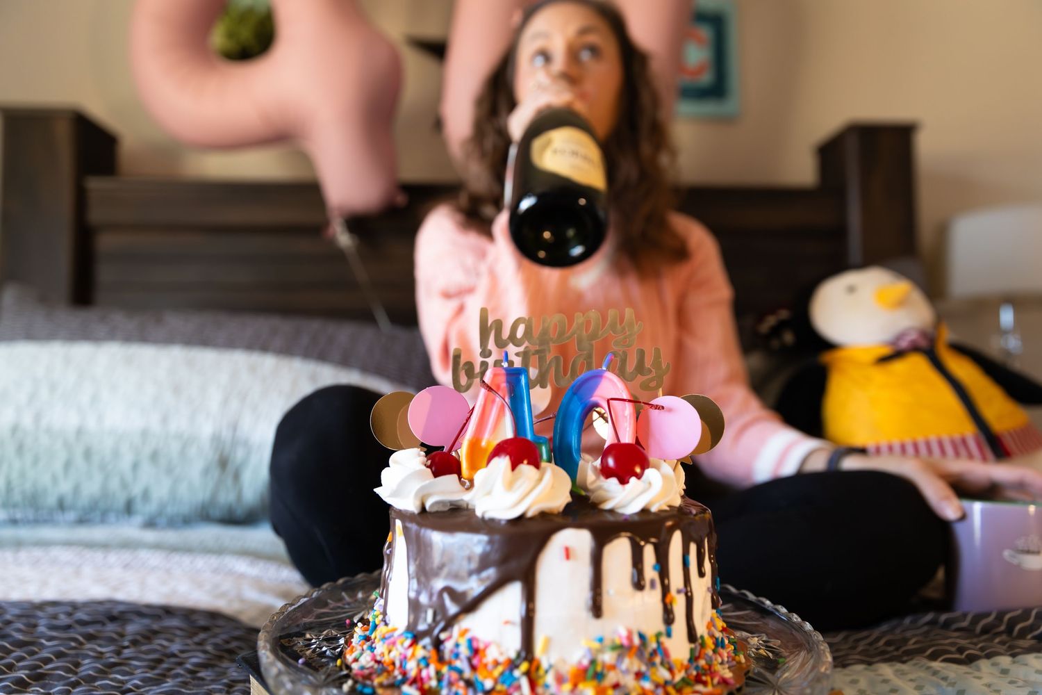 A chocolate drip birthday cake with colorful decorations sits on a bed while someone holds a bottle of champagne- Rome GA