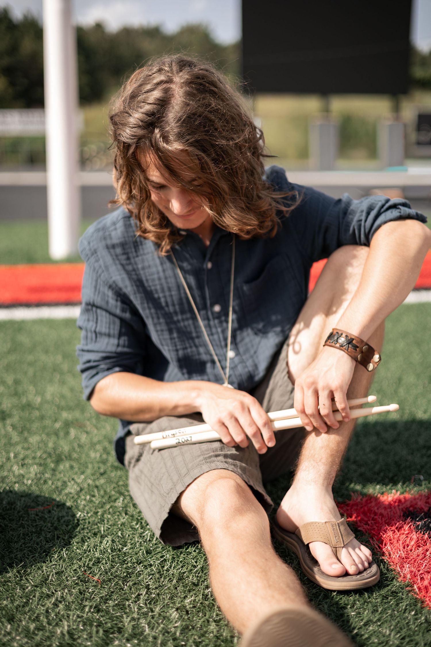 Young man with long hair sitting on grass holding drumsticks, wearing blue shirt and shorts outdoors. Senior Portraits Cedartown GA