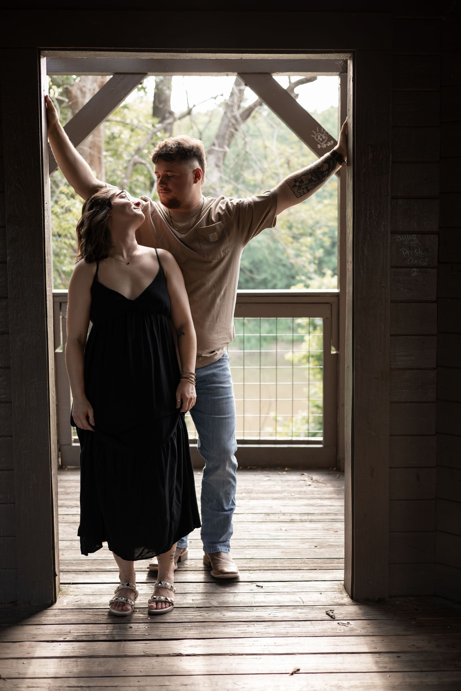 A couple stands in a rustic doorway, woman in black dress, man in jeans reaching up, with lush greenery behind them.