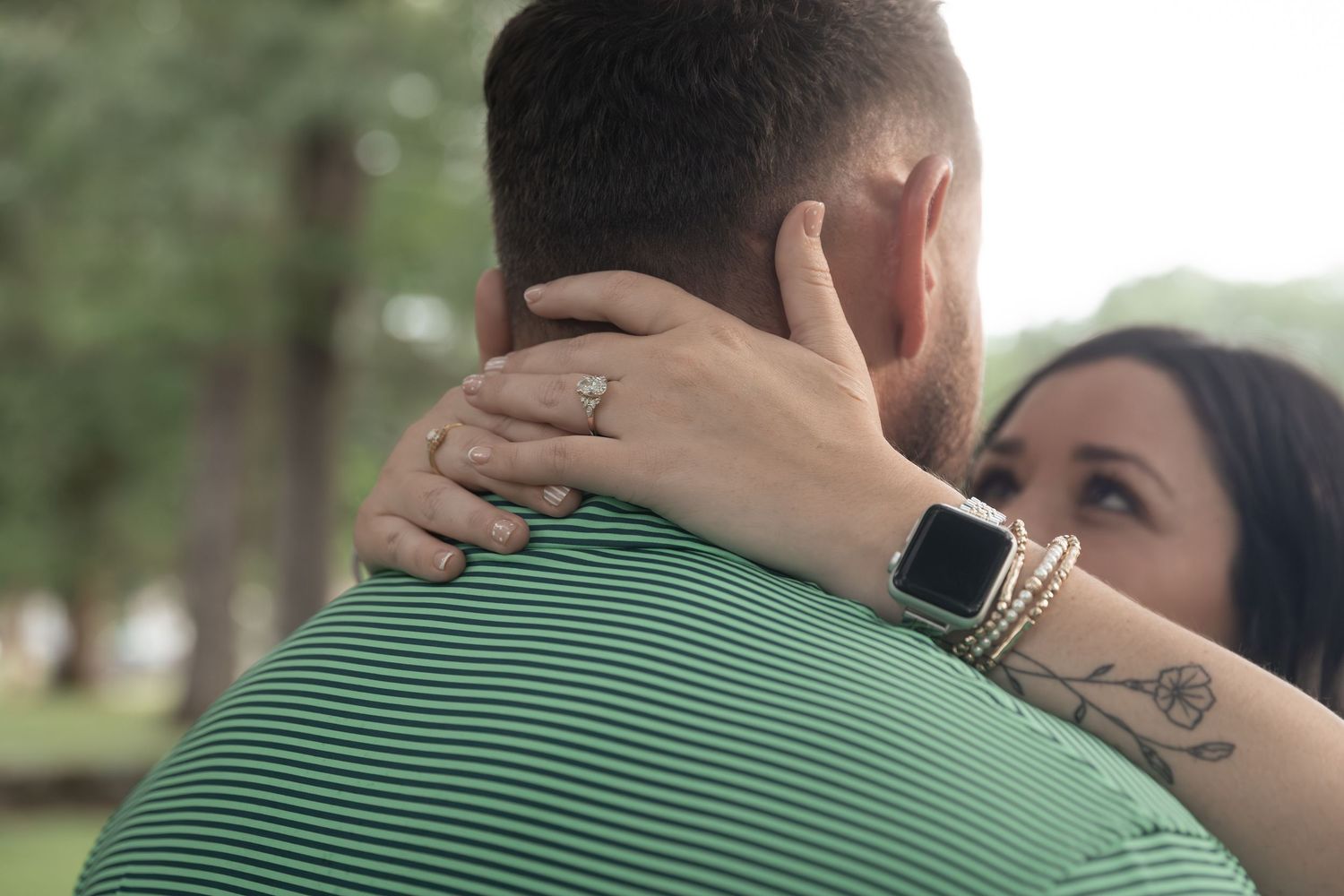 Couple embracing outdoors, woman wearing Apple Watch and ring with tattoo visible, man in green striped polo shirt.