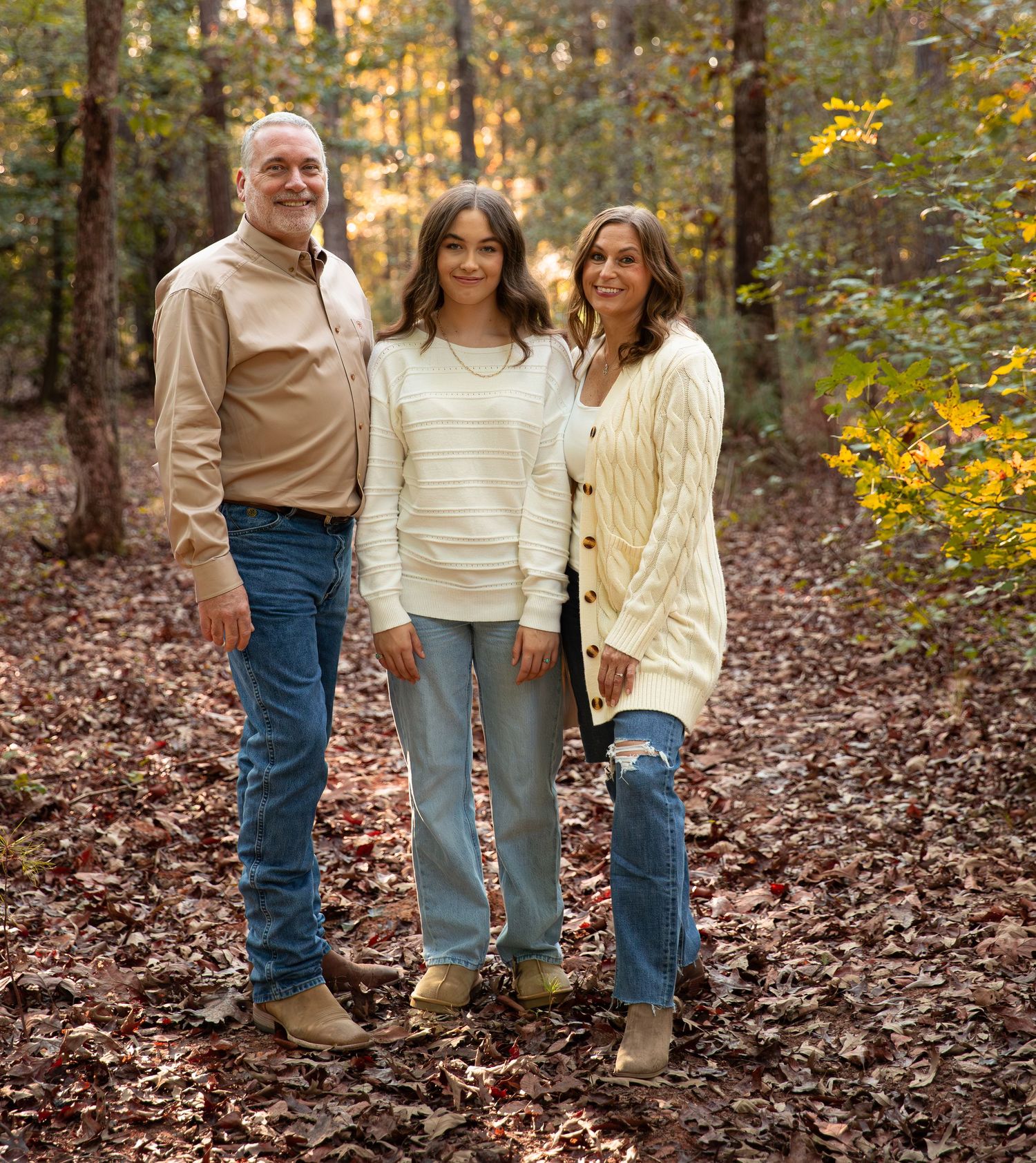 Family of three posing together in autumn forest with fallen leaves and colorful foliage backdrop.