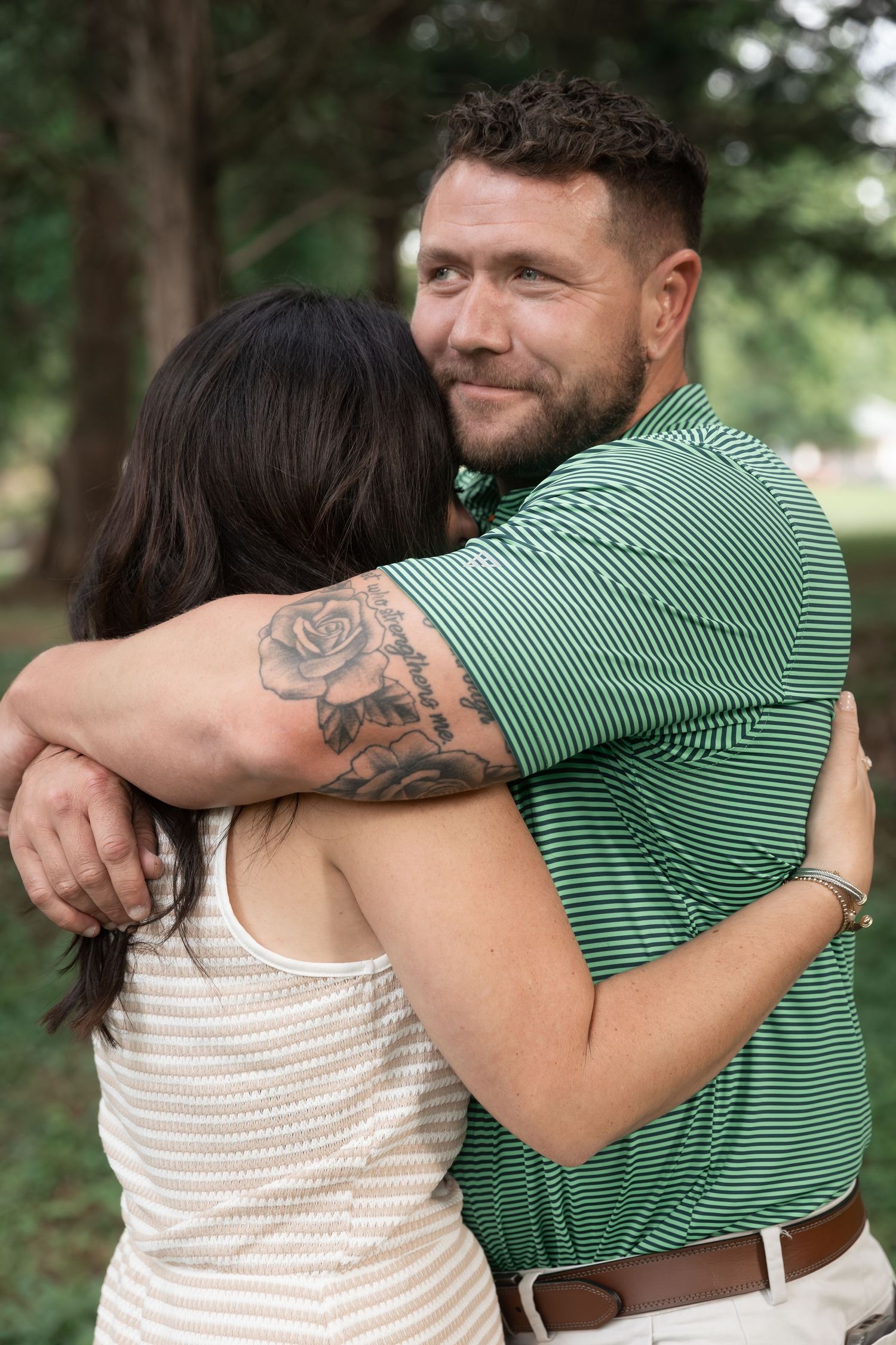 A man with a rose tattoo in a green striped polo embraces a dark-haired woman outdoors among trees. Cedartown, GA