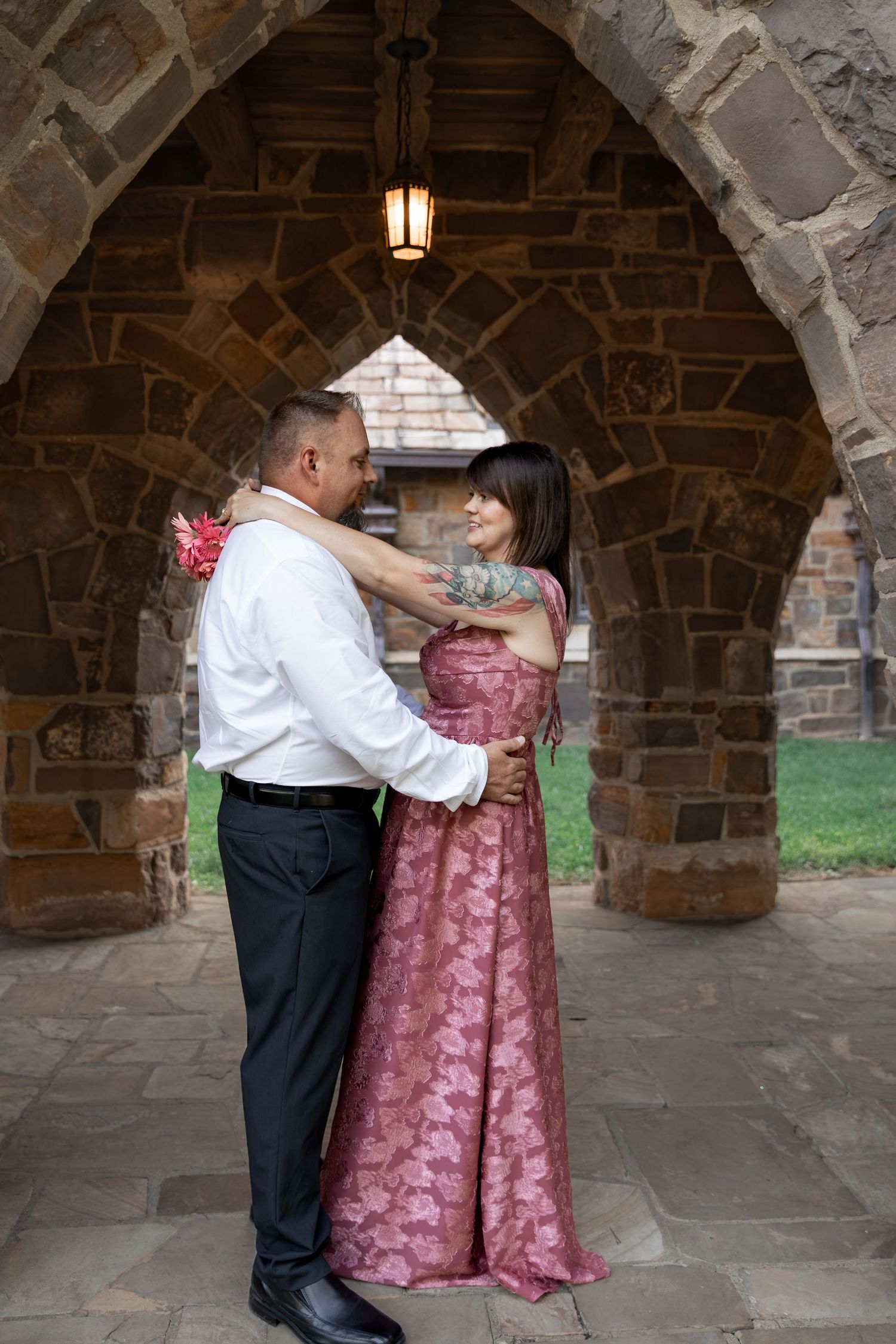Couple in formal attire embracing under a stone arch, woman in pink gown with tattoos, man in white shirt and black pants. Berry College