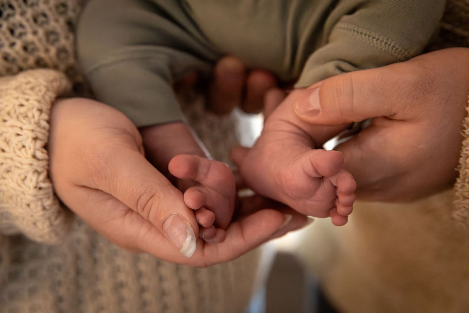 Adult hands gently cradle tiny newborn baby feet against a soft beige knit background.  Rome, GA Jessica M Cooper Photography LLC