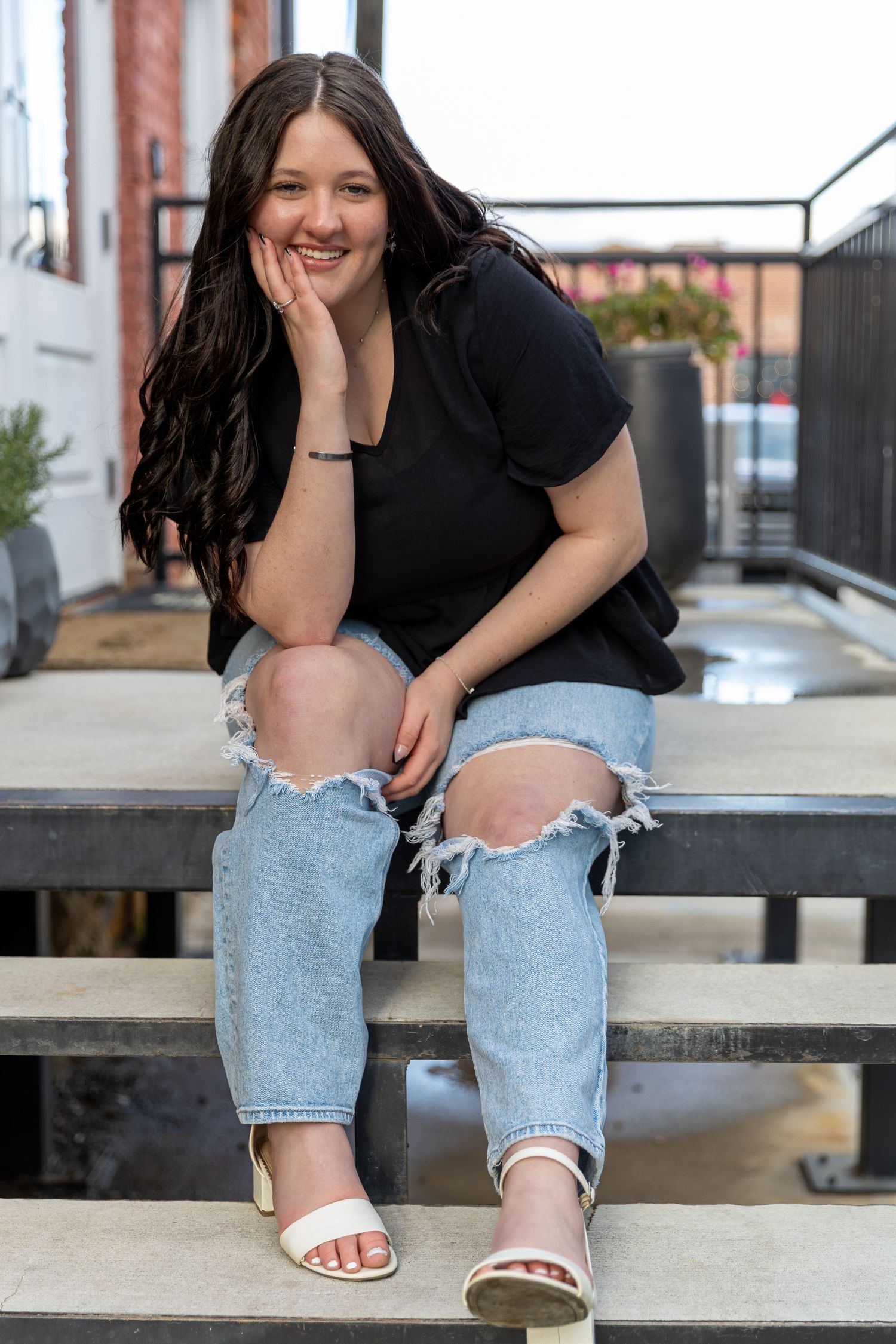 Young woman with dark hair smiling, wearing black tee, ripped jeans, and white heels, sitting on outdoor steps. Senior Portraits- GA