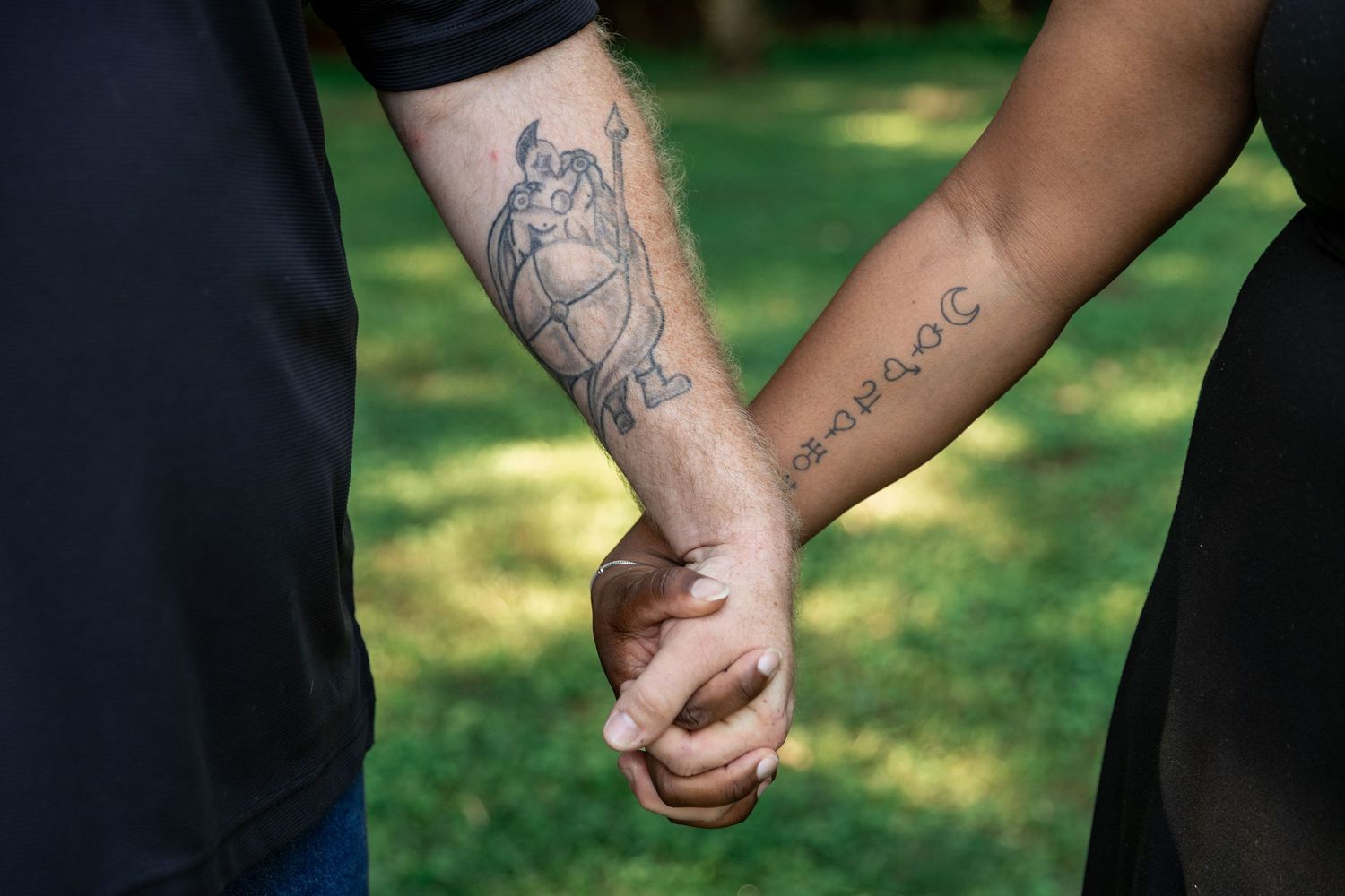 Two people holding hands outdoors, showing forearm tattoos, one with an angel design and one with script writing. Euharlee Covered Bridge