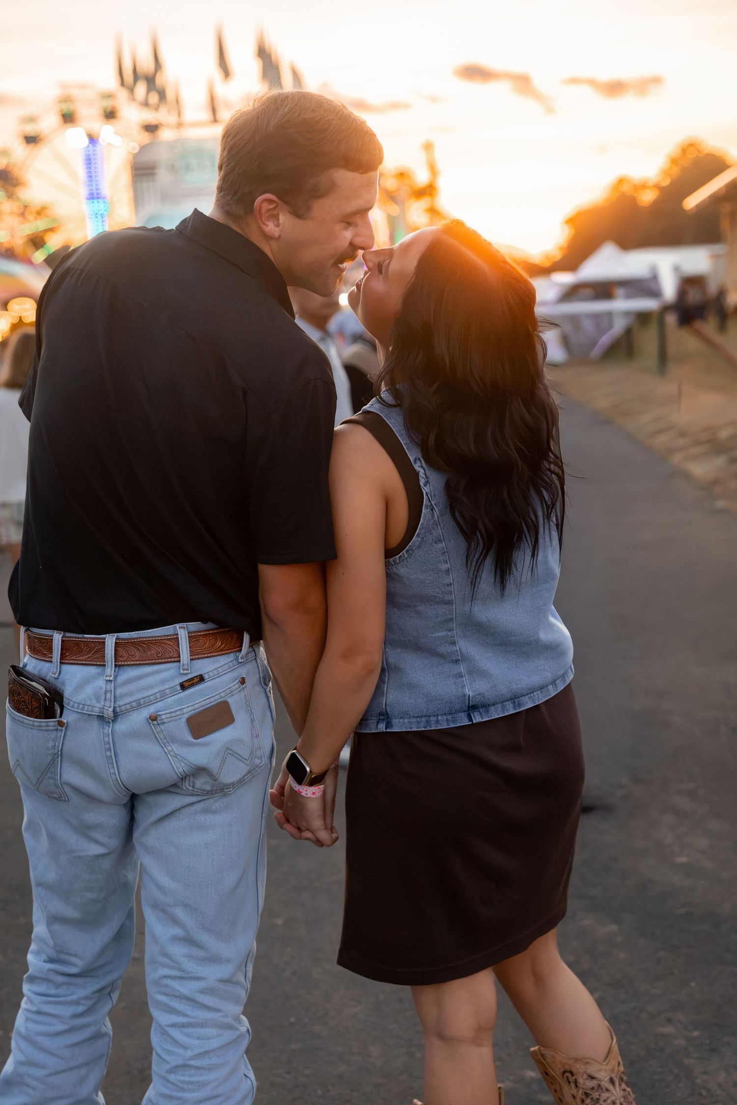 Couple holding hands and smiling at sunset outdoors, man in black shirt and jeans, woman in denim vest and skirt.