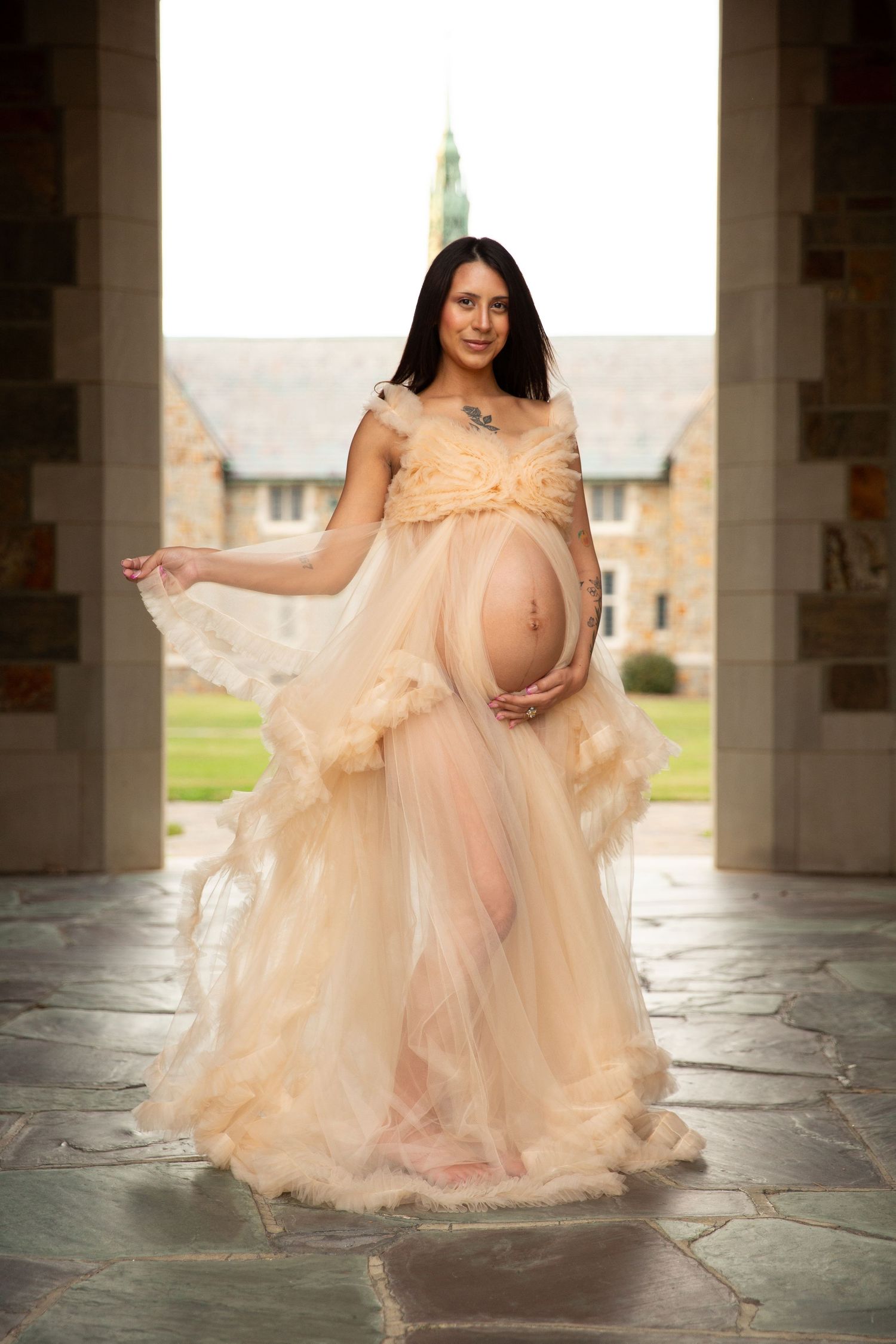 Pregnant woman in sheer peach tulle maternity gown posing outdoors at stone archway, cradling baby bump. Ford Buildings, Rome GA