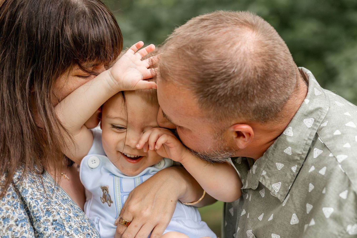 A joyful family moment with parents kissing and hugging a laughing baby outdoors in a lush green setting.