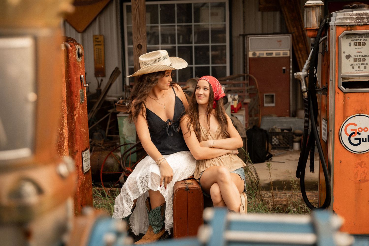 Two women in vintage outfits sit together near retro Gulf gas pumps at an old rustic station.