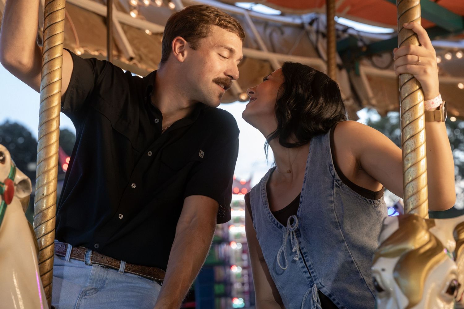 Romantic couple sharing a kiss on a colorful carousel at a sunny outdoor fair.