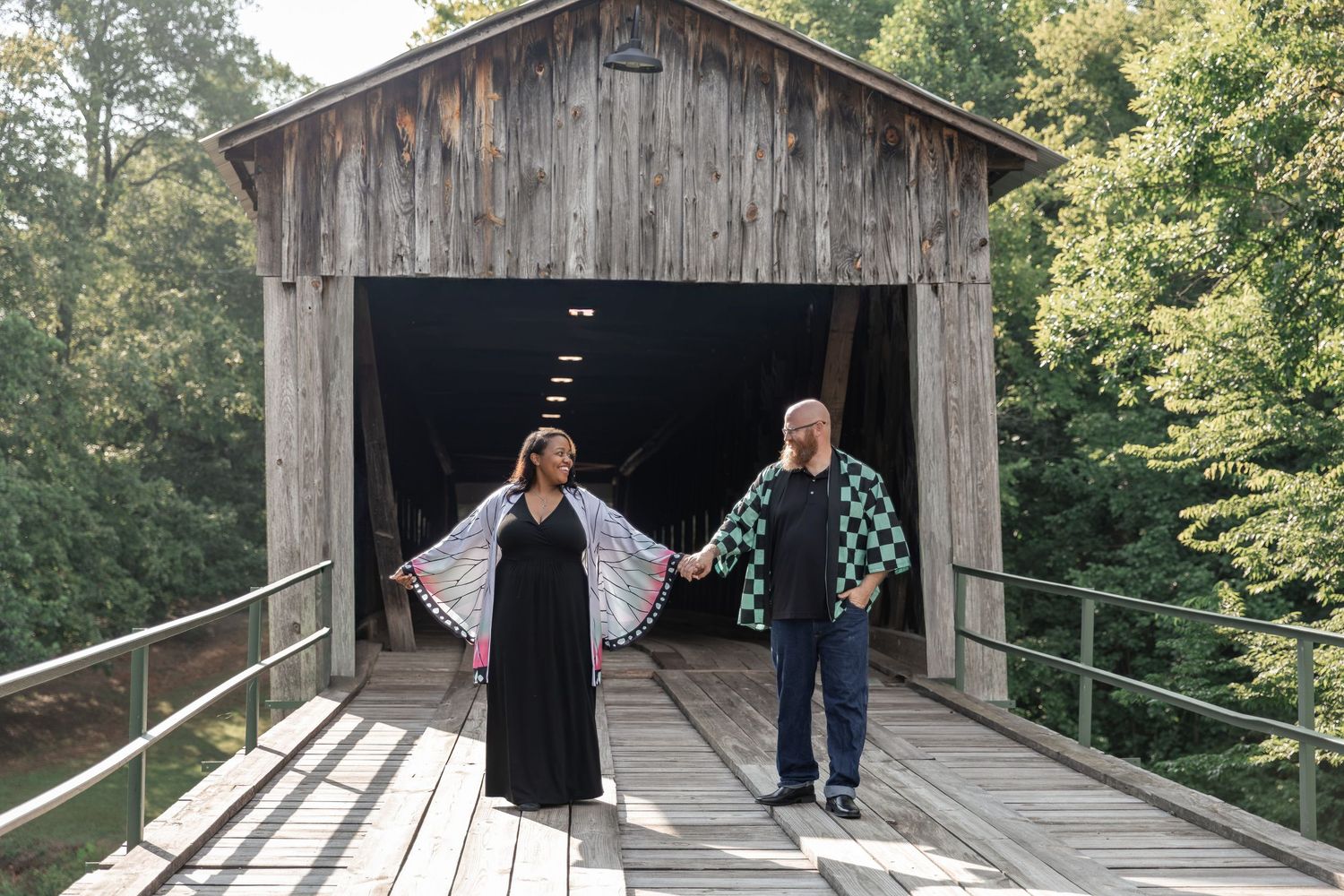 Couple posing at a rustic wooden covered bridge entrance surrounded by lush green trees. Euharlee Covered Bridge