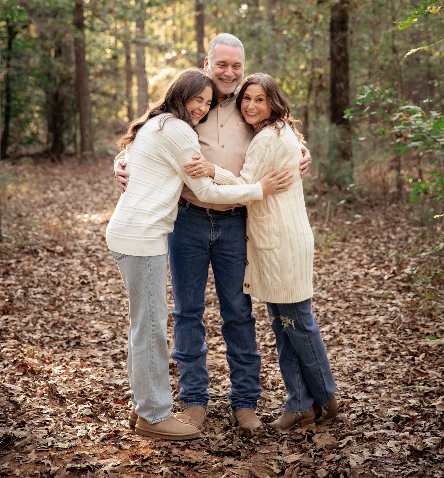 A man and two women share a warm hug outdoors in a wooded area covered with fallen autumn leaves.
