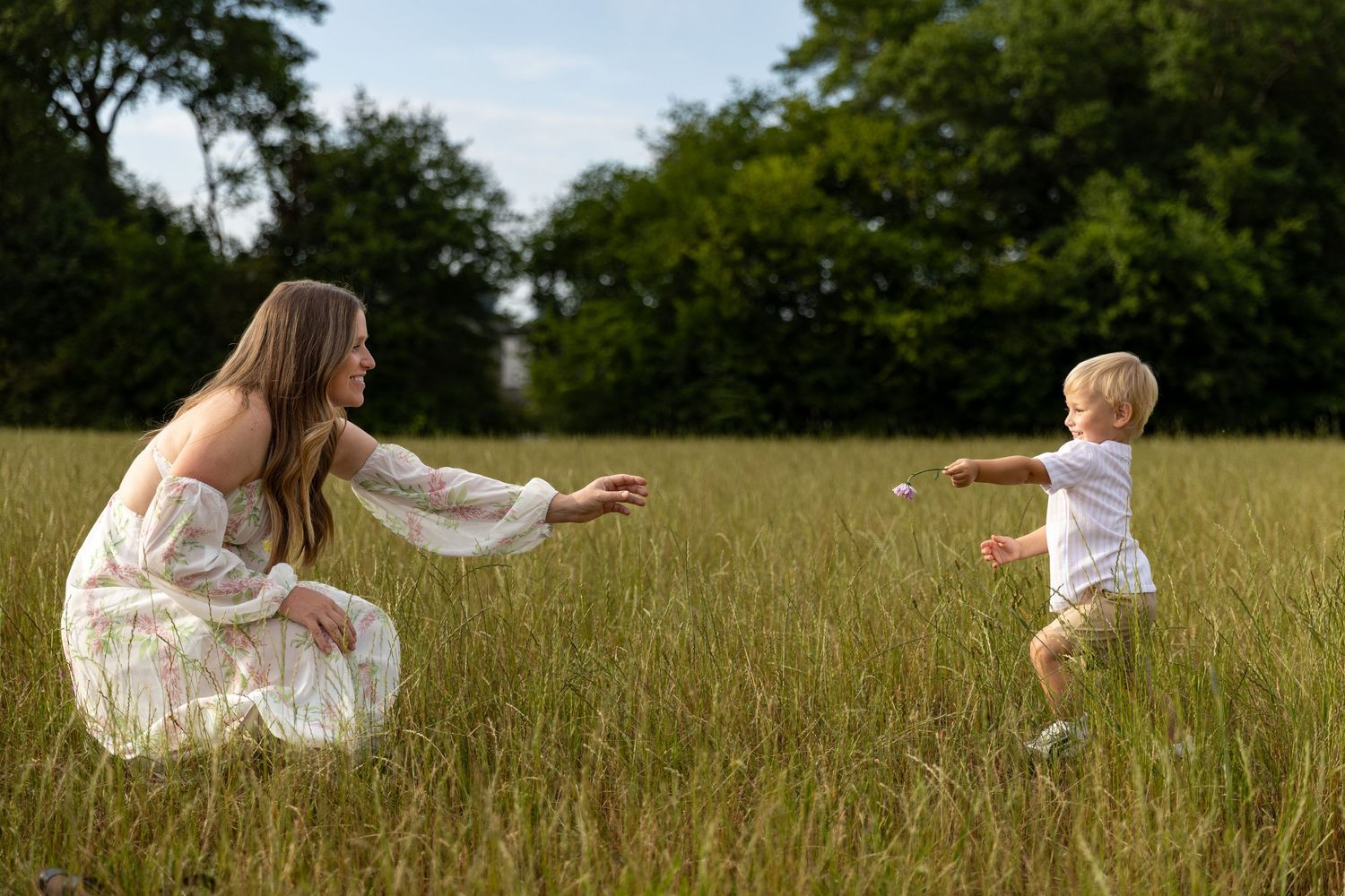 A mother in a white dress crouches in a green field, arms outstretched toward her toddler running to her.