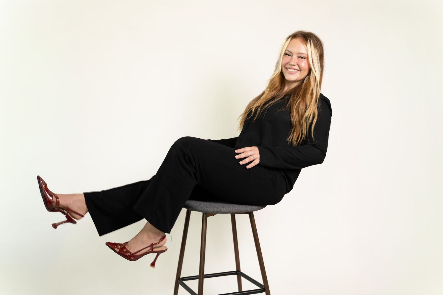 Smiling blonde woman in black pantsuit sitting casually on a barstool with legs raised, wearing brown heels. Branding session Acworth, GA