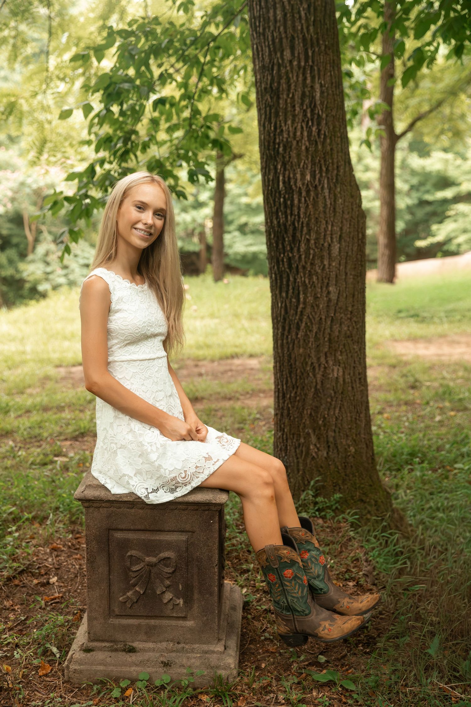 Smiling blonde girl in white lace dress and cowboy boots sitting on stone pedestal in lush green park.