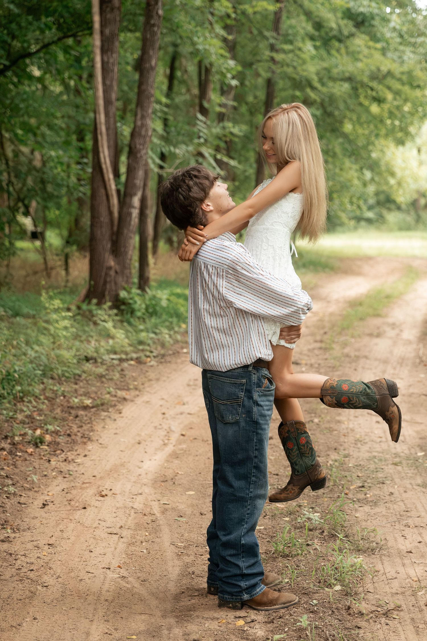 A young couple embraces on a rustic dirt path surrounded by lush green trees, as the man lifts the woman in cowboy boots.