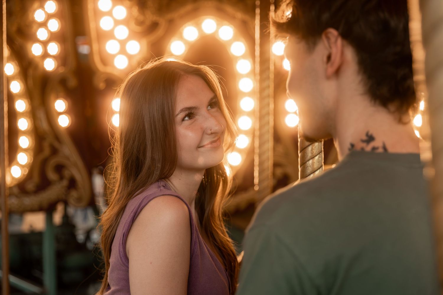 A young woman smiles warmly at a man near a glowing carousel at a fairground.