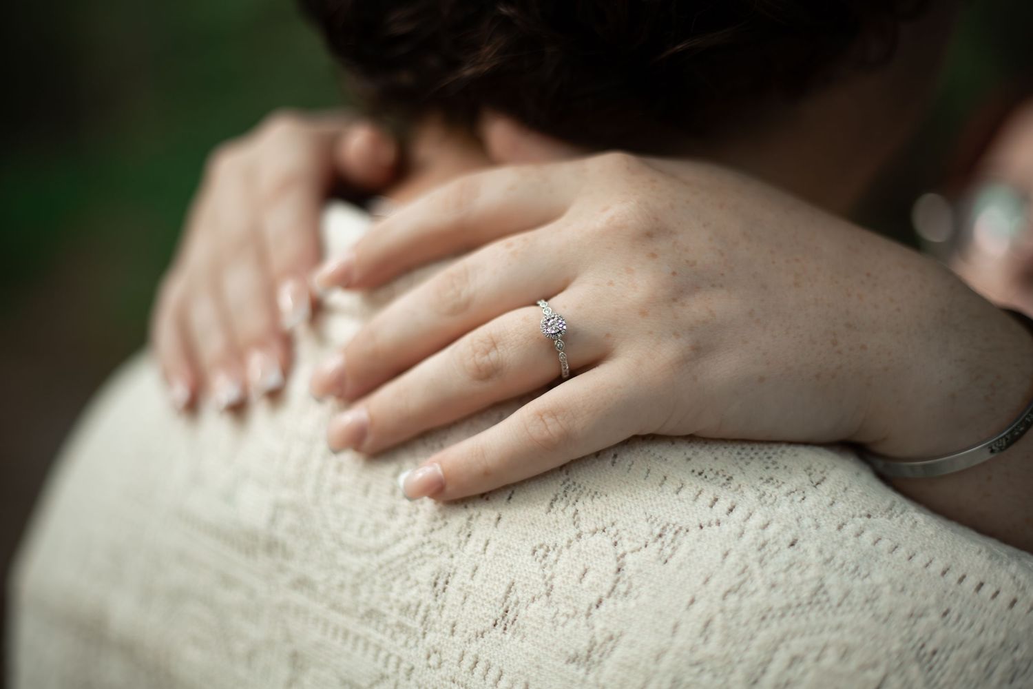 Close-up of a woman's hand with a diamond engagement ring resting on a partner's shoulder in a lace dress.