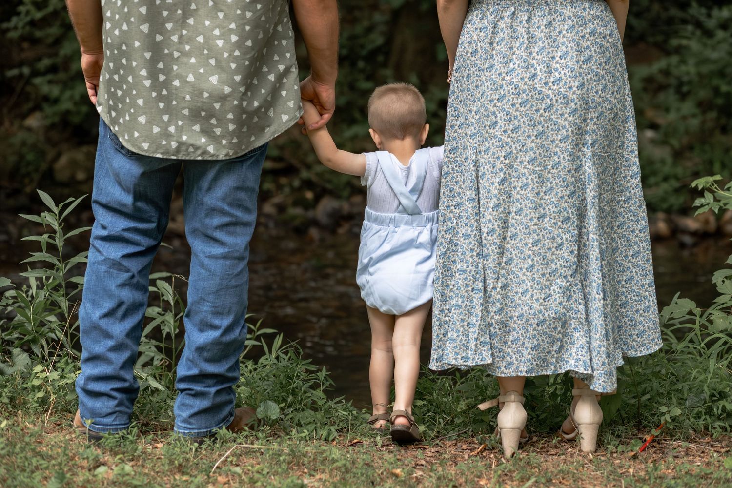 A toddler walks hand-in-hand with parents through a lush green garden path in a heartwarming family moment.