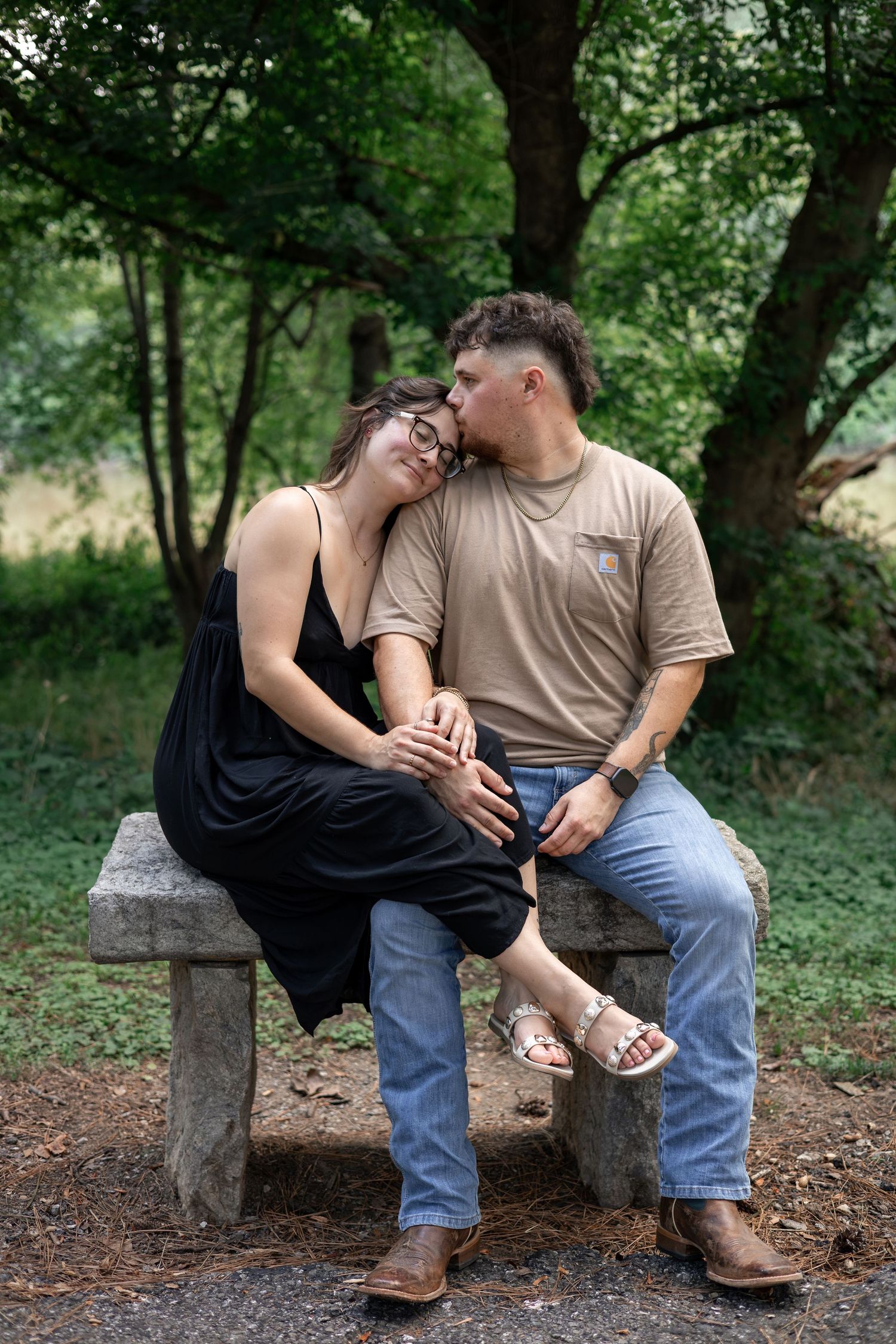 A couple sits on a stone bench outdoors, the man kissing the woman's head as she leans on his shoulder.