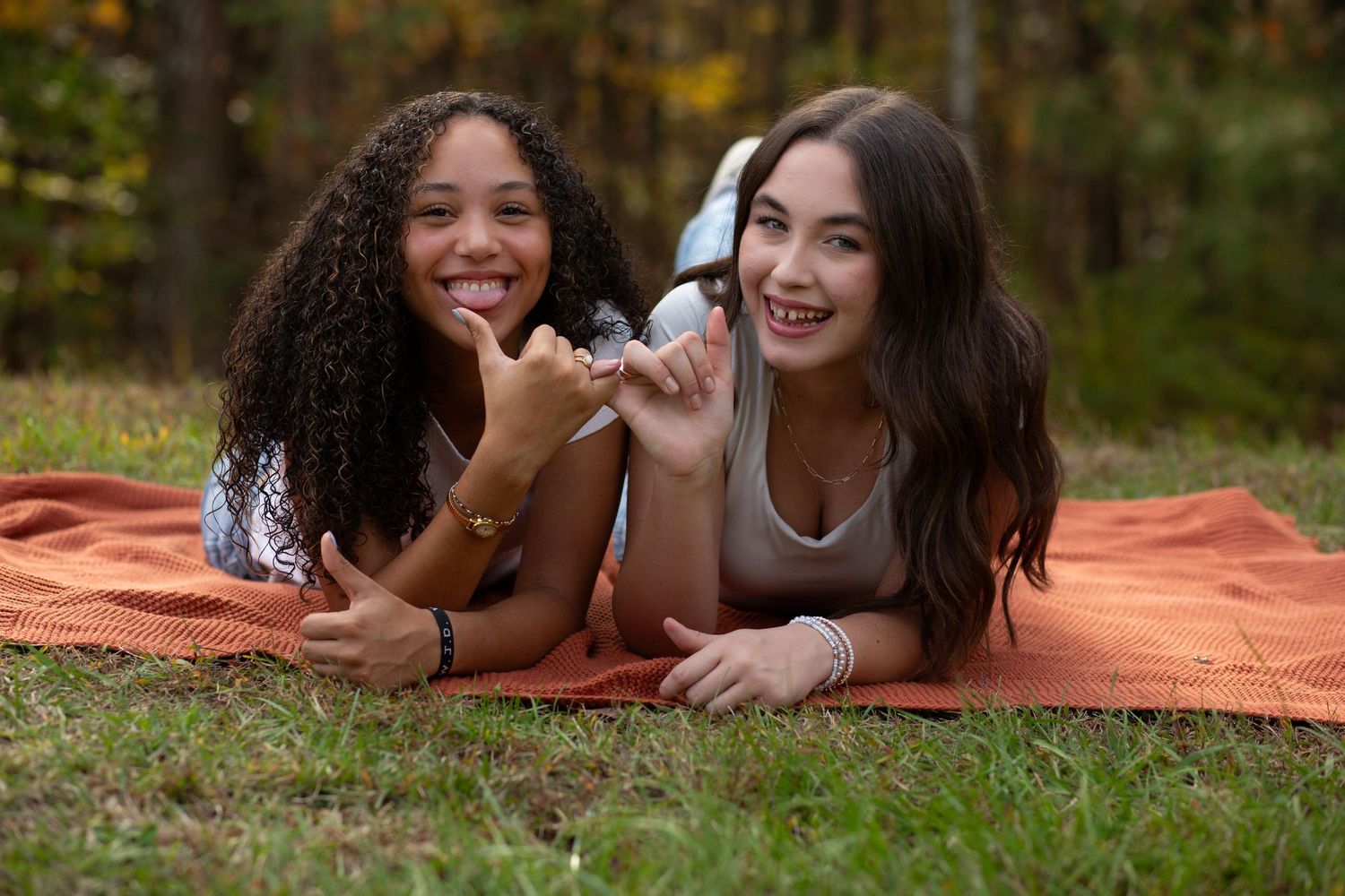 Two smiling teenage girls lying on an orange blanket outdoors in a park with autumn trees in the background.